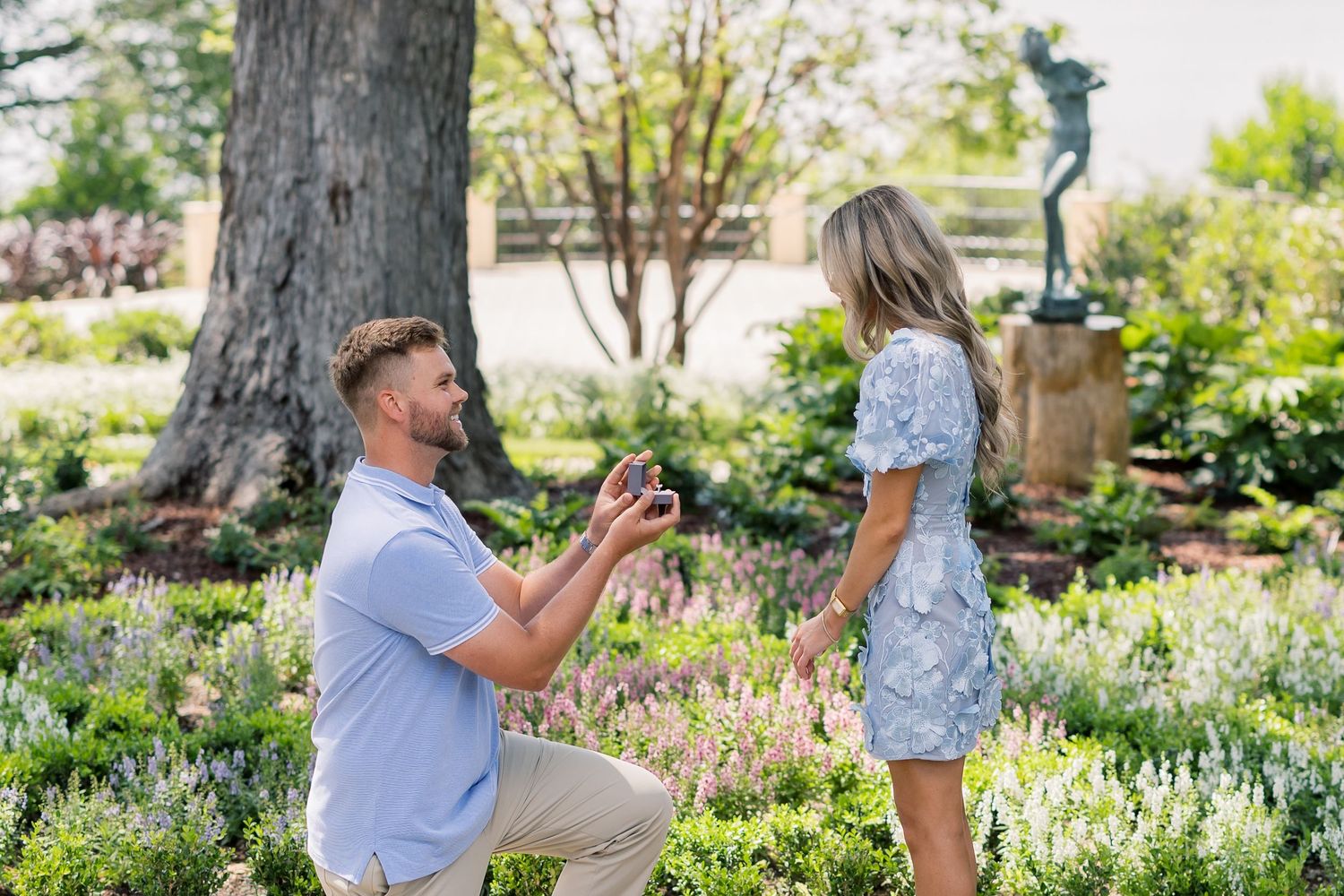 Man kneeling on one knee proposing to woman in a lush garden with flowers and a bronze statue.