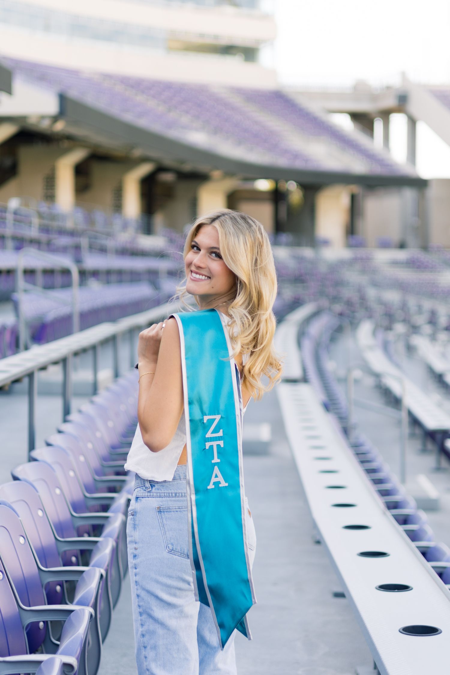 Smiling blonde woman wearing a teal ZTA graduation sash poses in purple stadium bleachers.
