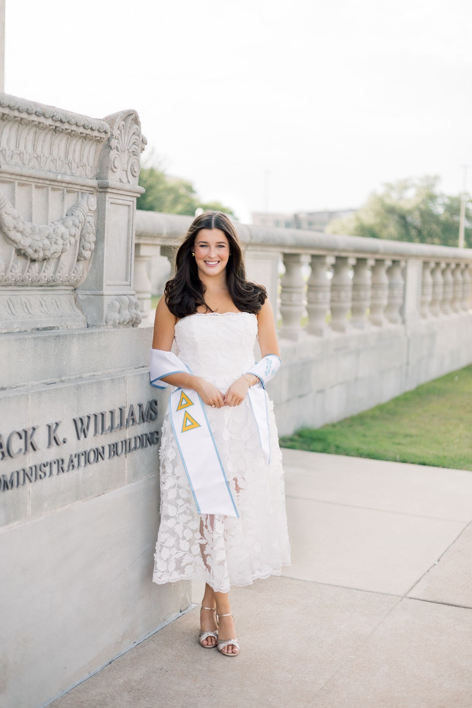 Brunette graduate in white strapless lace dress holding Delta Delta Delta sorority stole outdoors.