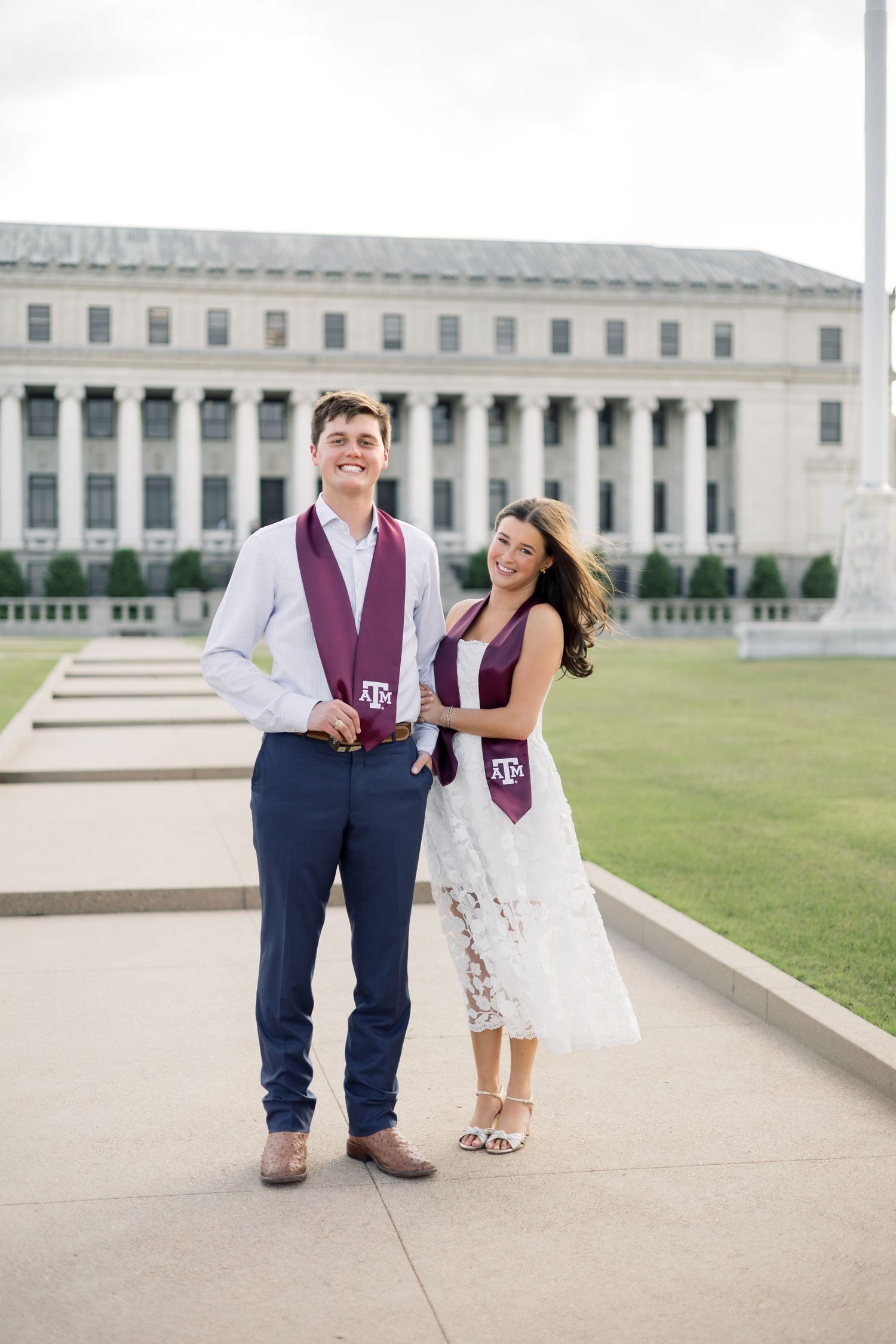 Smiling couple standing outdoors wearing matching Texas A&M maroon graduation stoles in front of campus building.