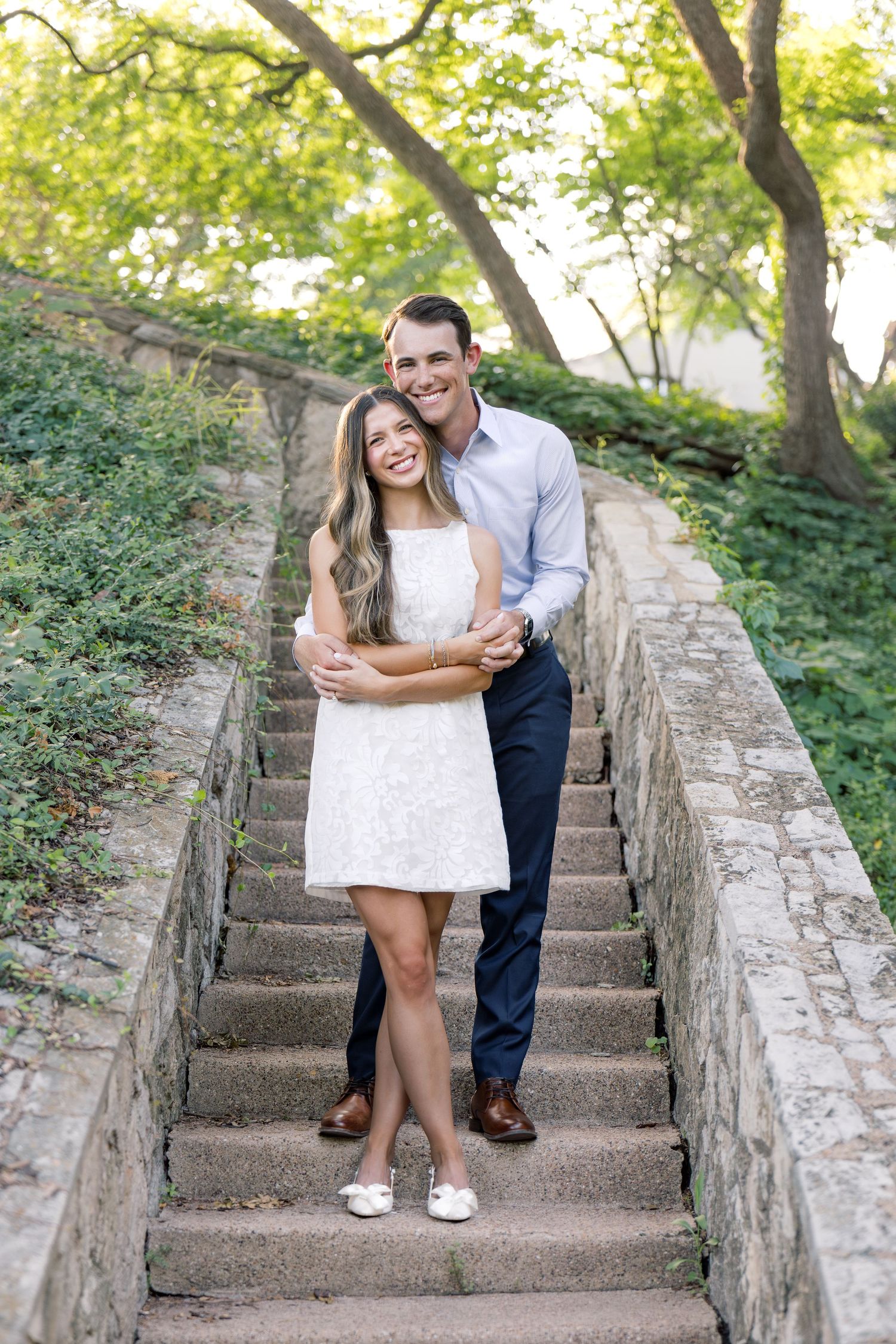 Couple posing on stone steps outdoors, woman in white dress, man in blue shirt and navy pants, surrounded by greenery.