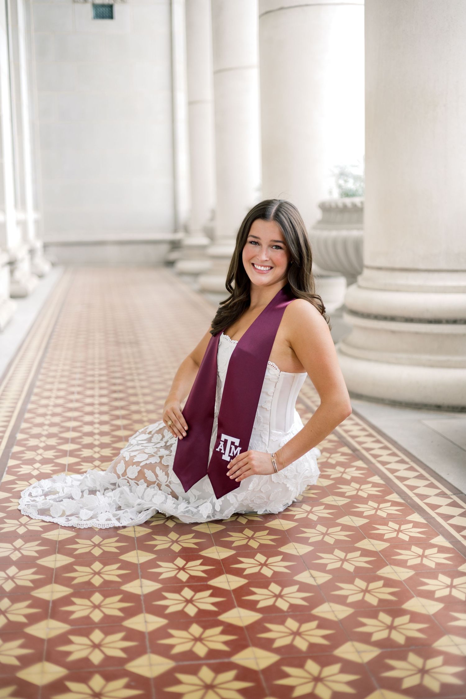Smiling woman in white strapless lace dress with Texas A&M graduation stole seated on decorative tiled floor by columns.