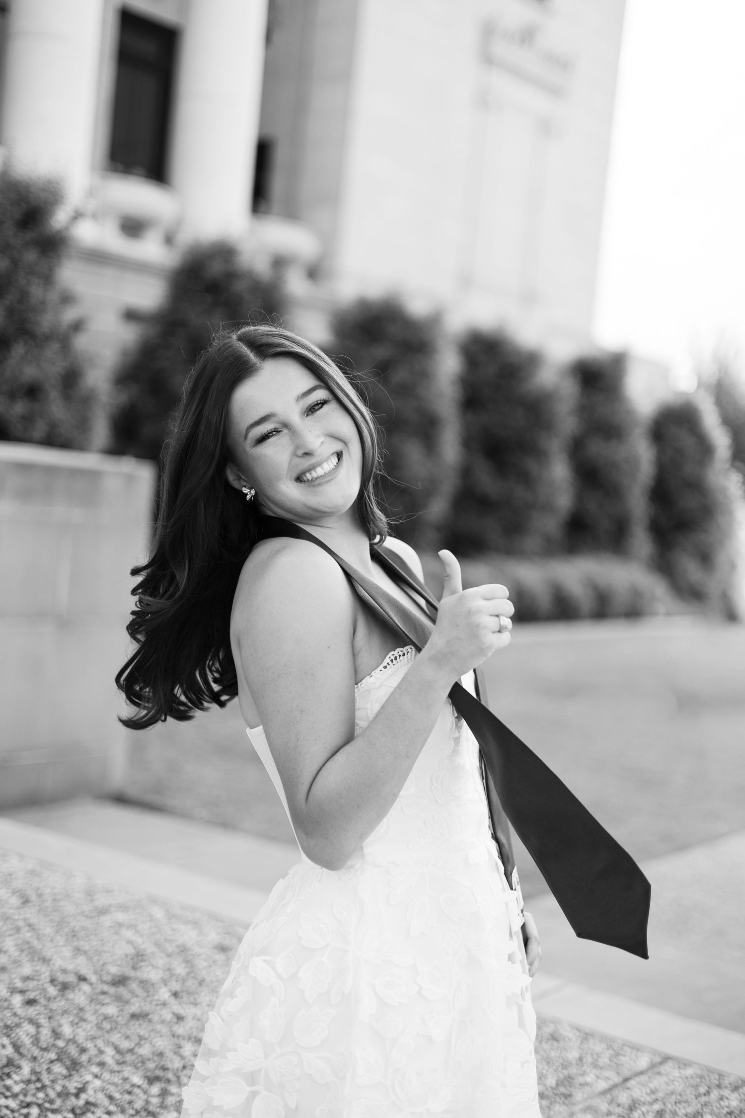 Young woman in white lace dress smiling over shoulder holding graduation stole in black and white photo.