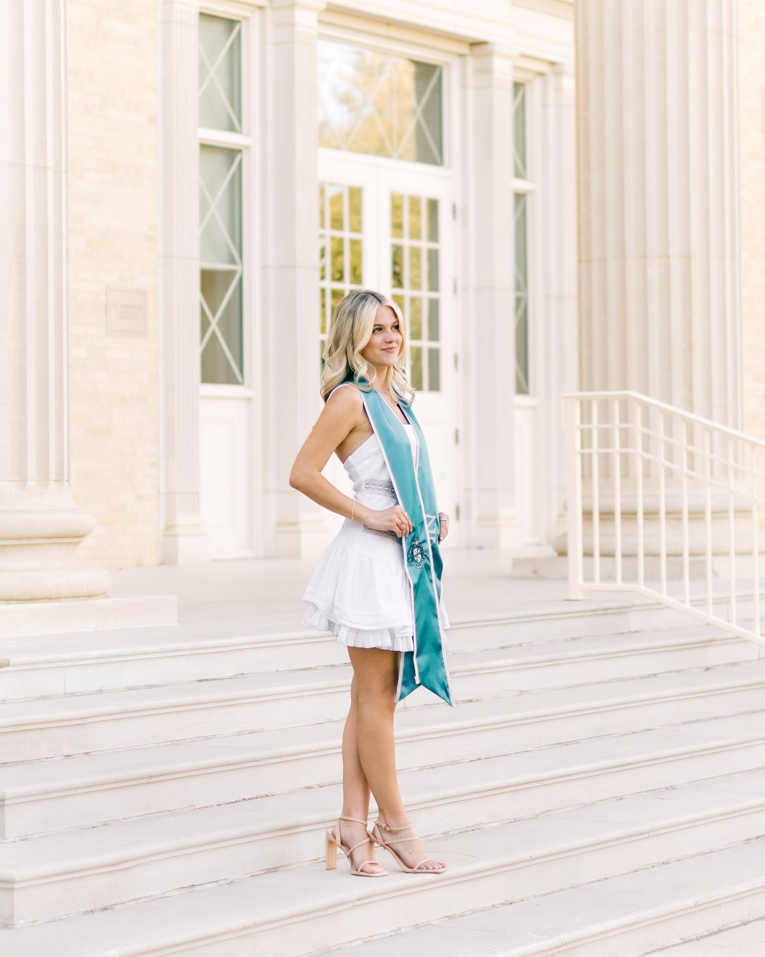 Blonde graduate in white dress and teal sash standing on steps of neoclassical university building.
