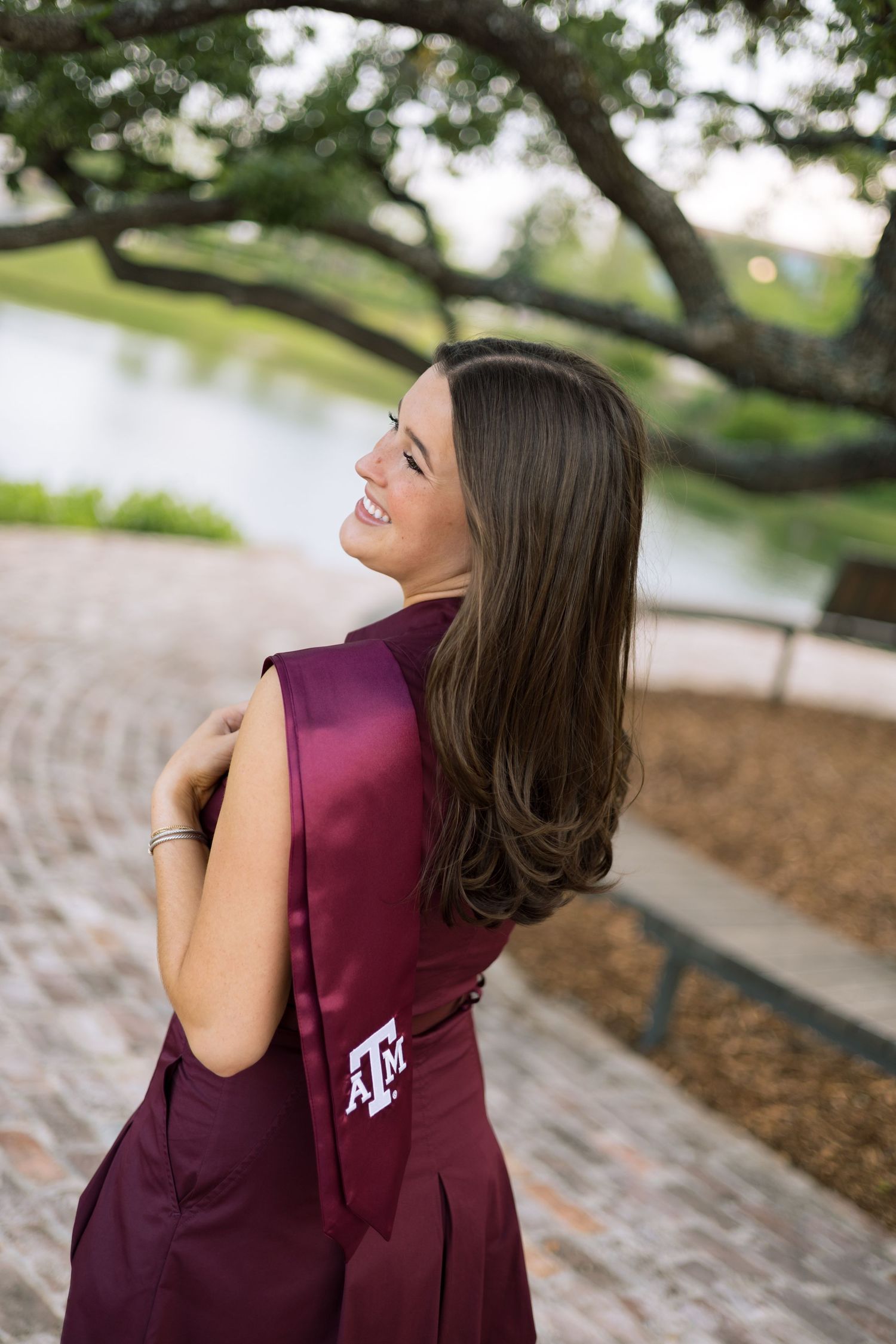 Brunette woman in maroon Texas A&M graduation dress laughing outdoors near a waterway.