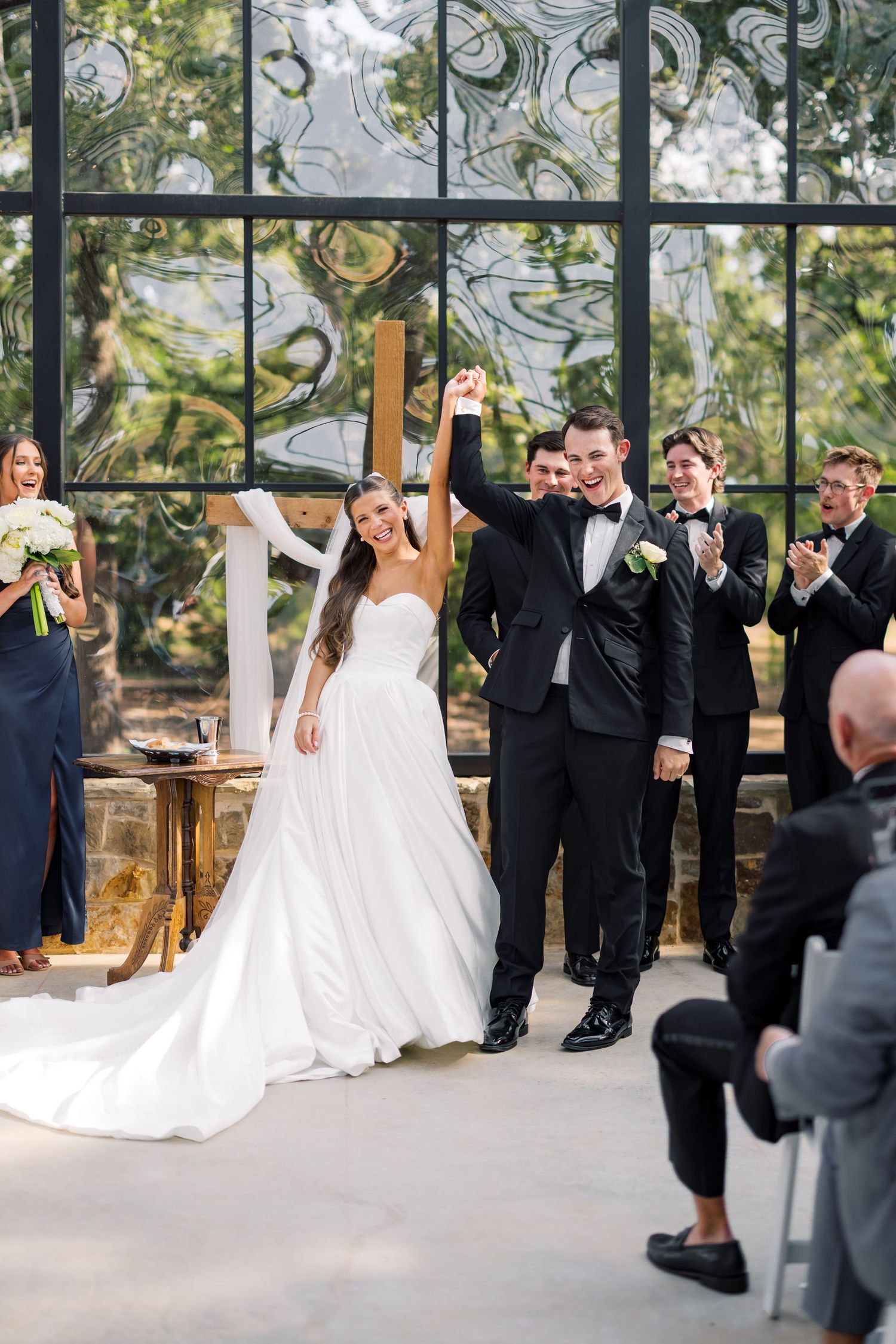 Bride and groom raise hands in celebration at elegant indoor wedding ceremony with bridal party applauding behind them.