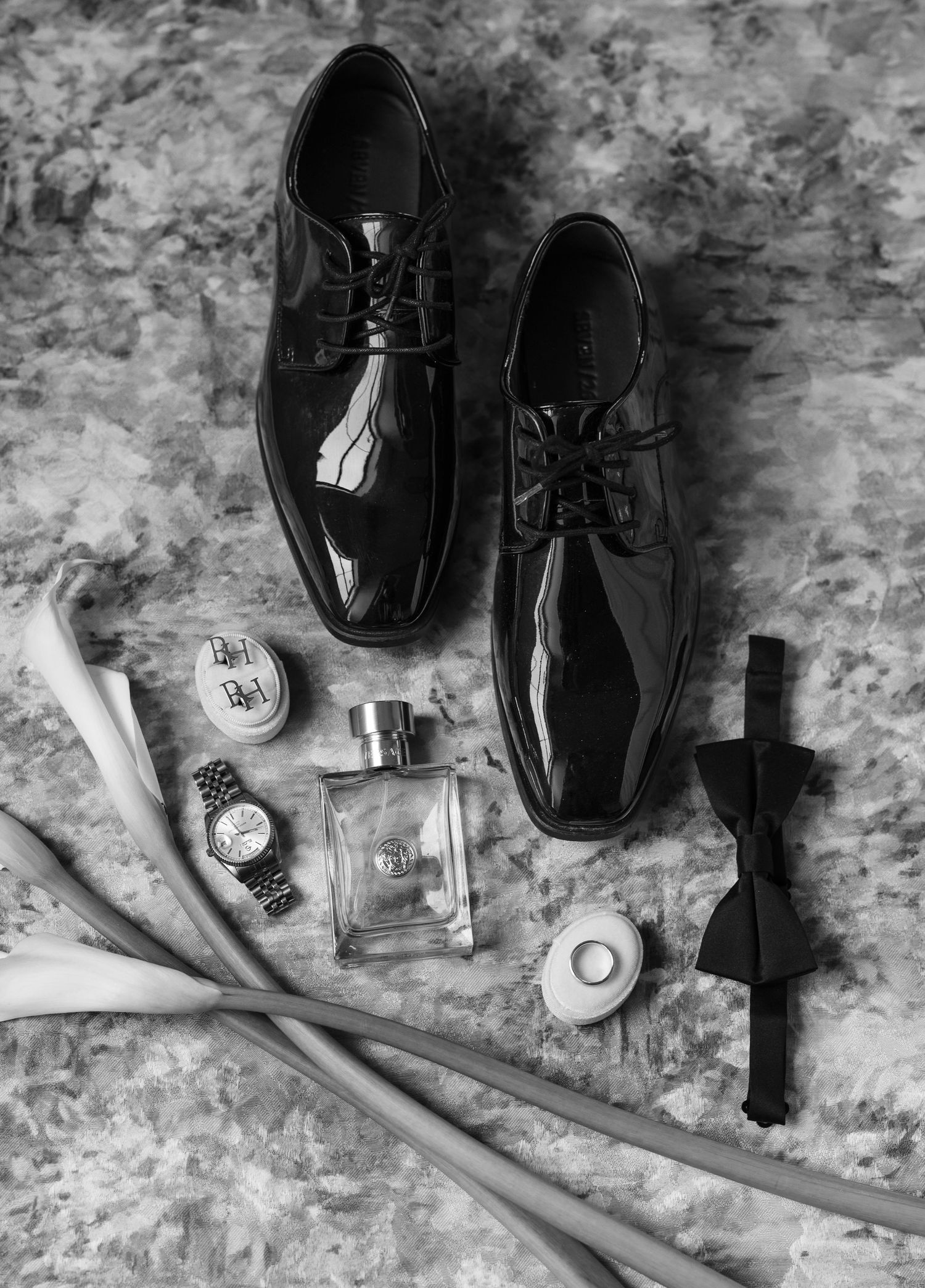 Black and white flat lay of mens wedding accessories including patent leather shoes, bow tie, watch, cologne, and ring.