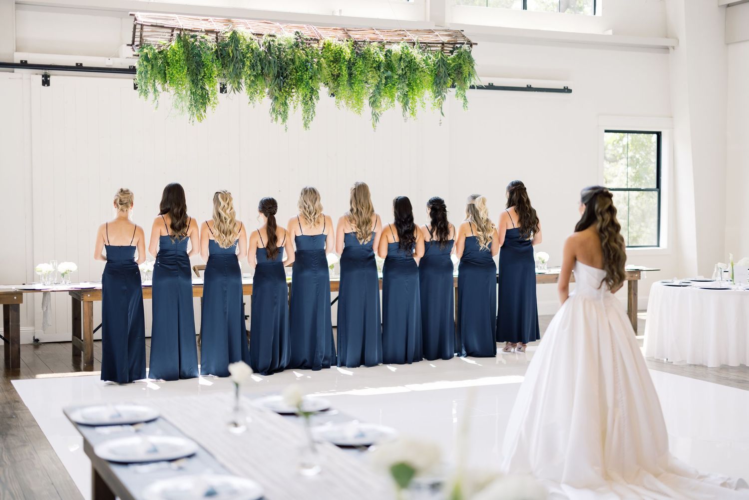 Bride reveals dress to bridesmaids in navy blue gowns standing beneath a lush greenery installation in an elegant venue.