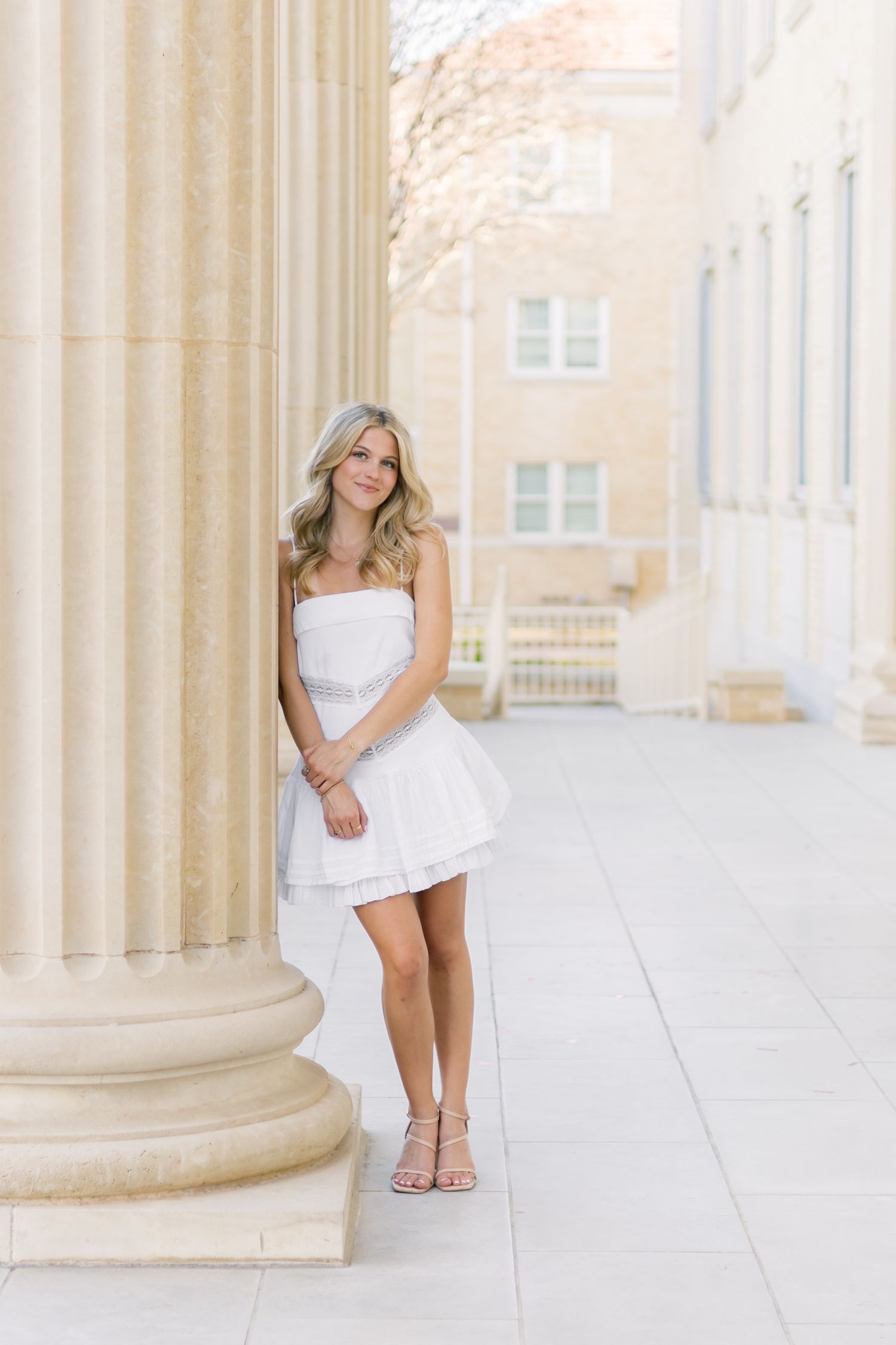 Blonde woman in white strapless mini dress leaning against a large stone column on a campus.