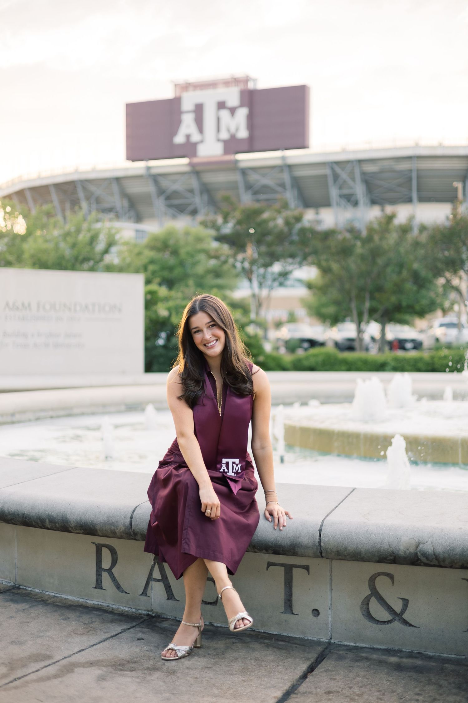 Texas A&M graduate in maroon dress sitting by fountain with Kyle Field stadium backdrop.
