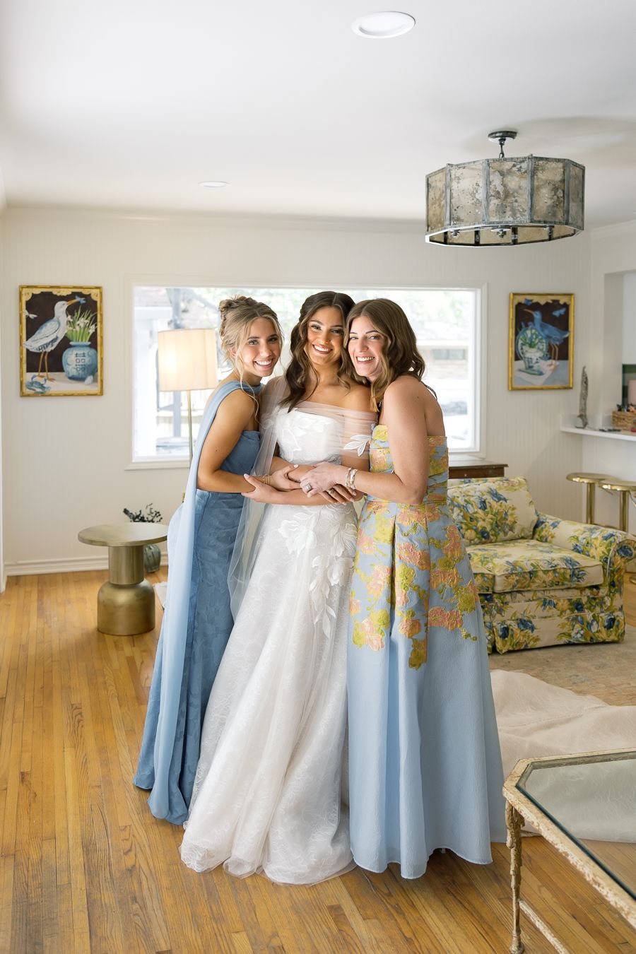 Bride in white gown poses with two bridesmaids in blue dresses inside an elegantly decorated room.