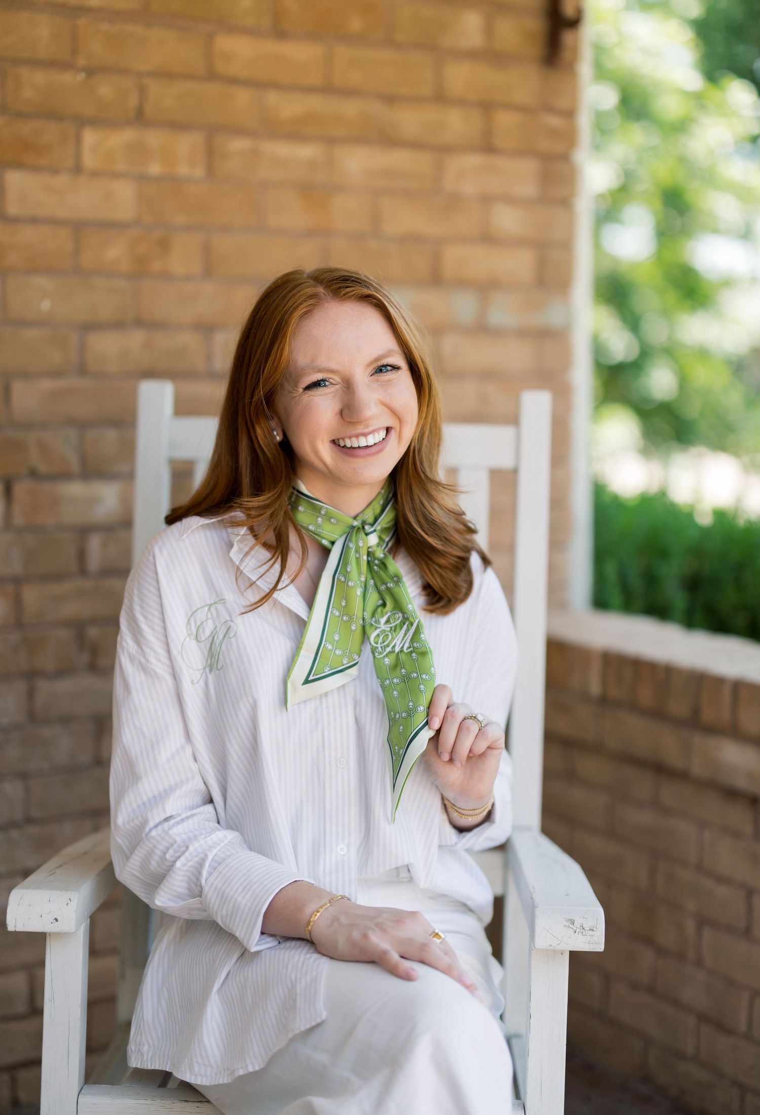 Smiling redhead woman in white outfit with green scarf sits in white rocking chair on brick porch.