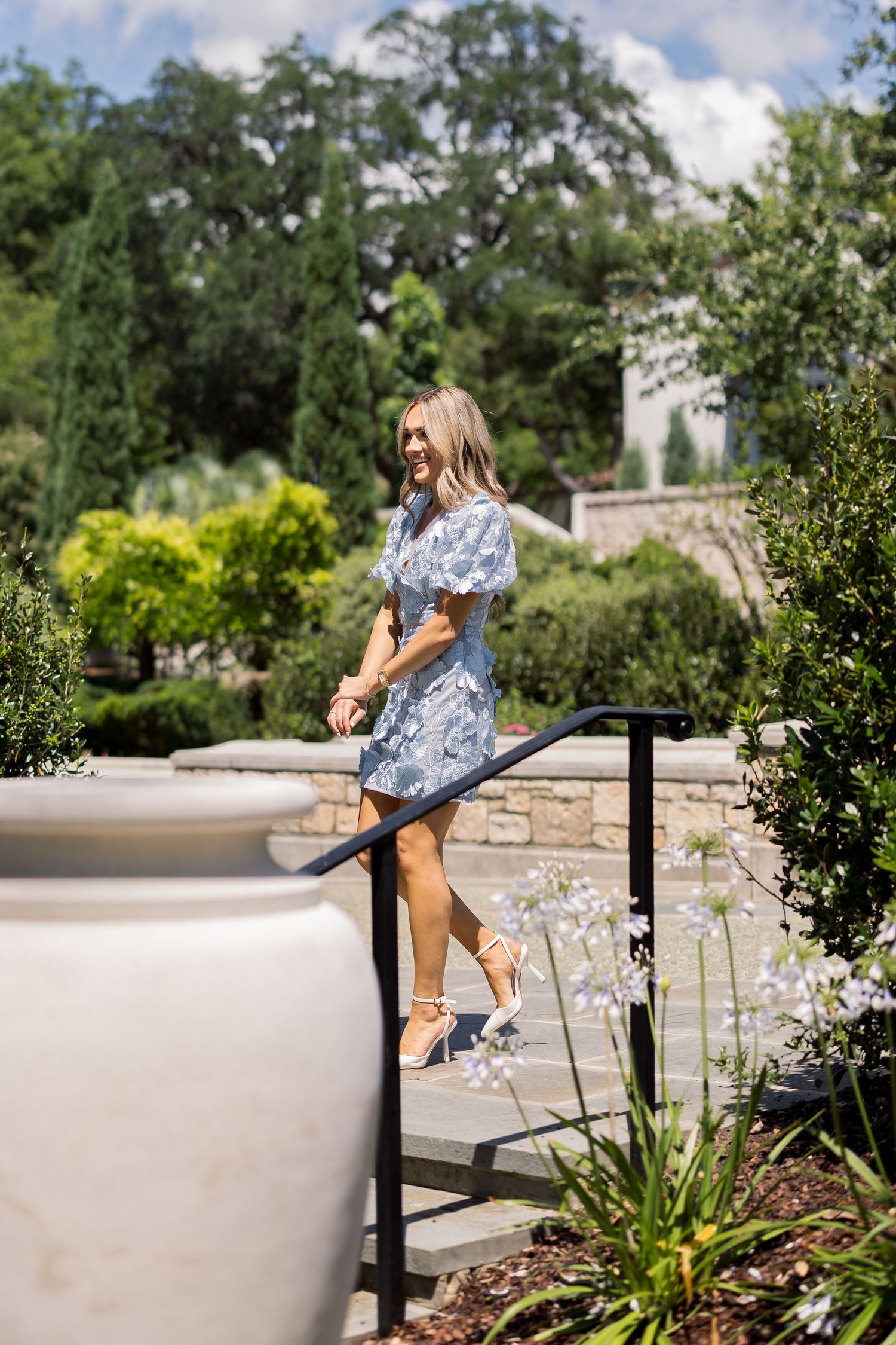 Woman in blue floral mini dress and white heels smiling on garden steps surrounded by lush greenery.