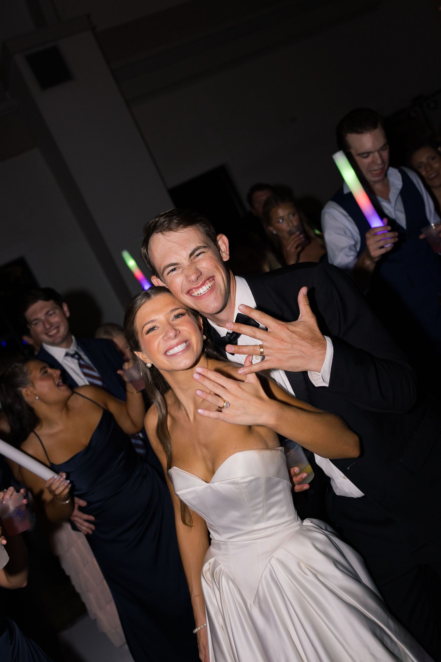 Joyful newlyweds dancing at wedding reception, bride in white strapless gown and groom in black tuxedo showing rings.