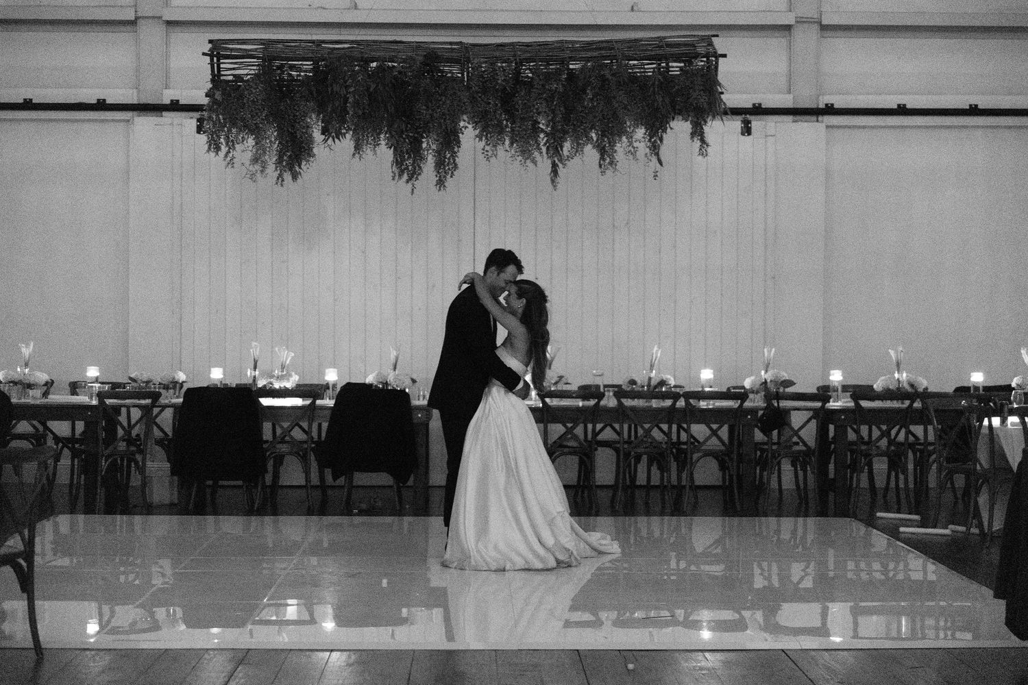 Black and white photo of bride and groom sharing first dance on reflective floor with candlelit reception tables behind them.