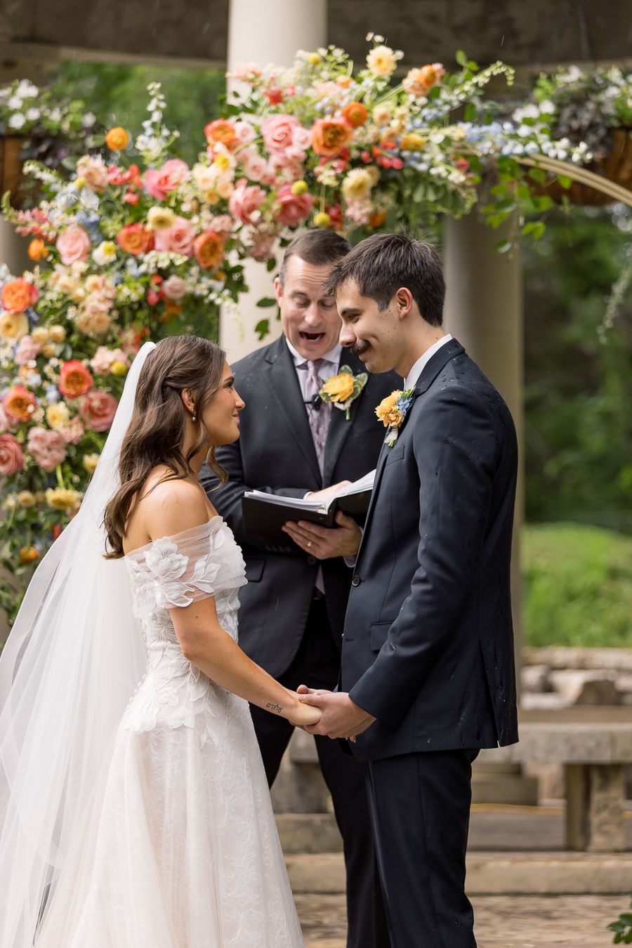 Bride and groom holding hands and laughing during outdoor wedding ceremony with floral arch.