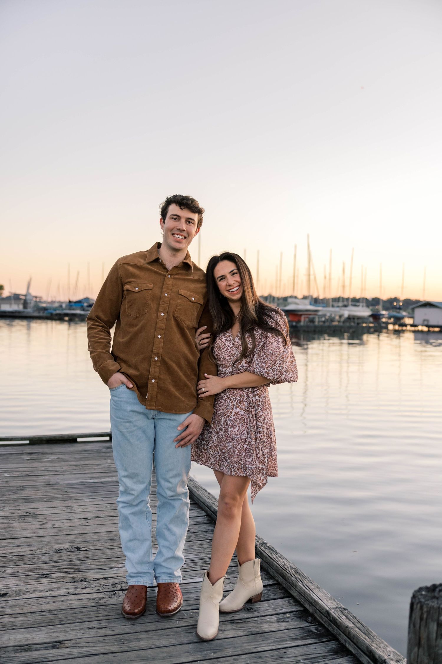 Happy couple posing on a wooden dock at sunset with sailboats reflected in calm harbor waters.