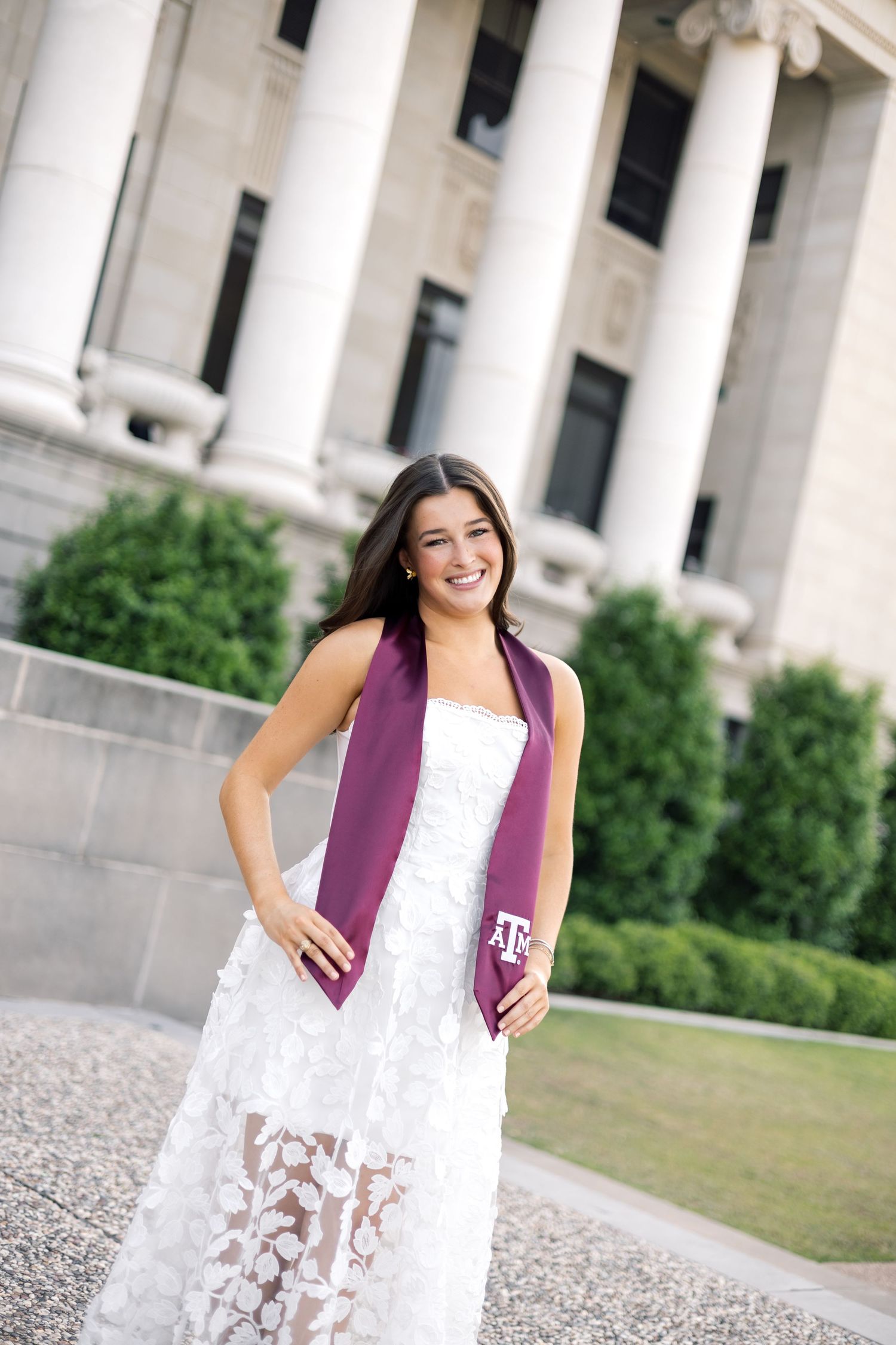 Smiling graduate in white lace dress wearing maroon Texas A&M stole outside Jack K. Williams building.