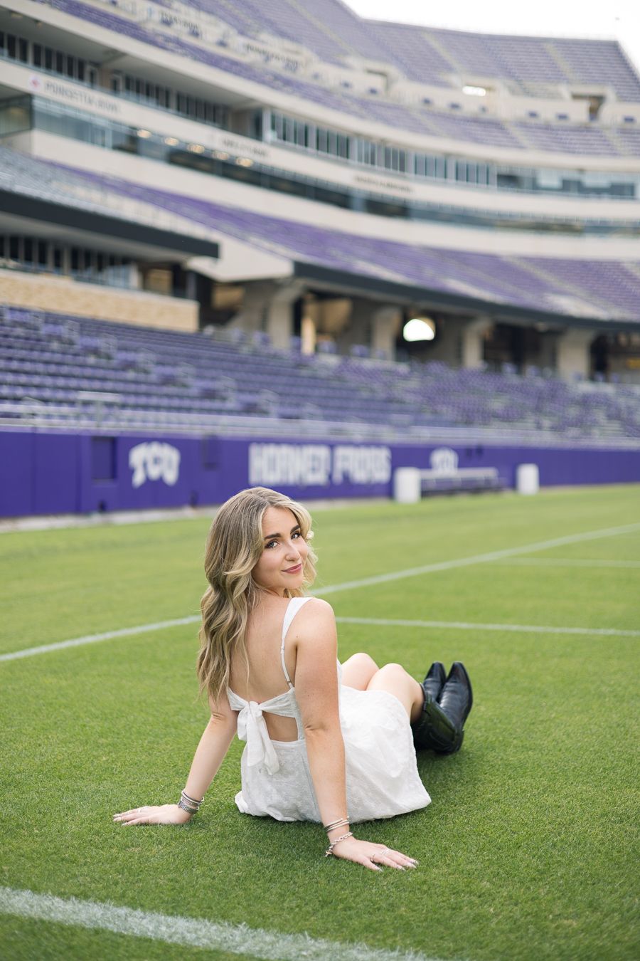 Young woman in white dress sitting on TCU Horned Frogs football field turf with purple stadium seats in background.
