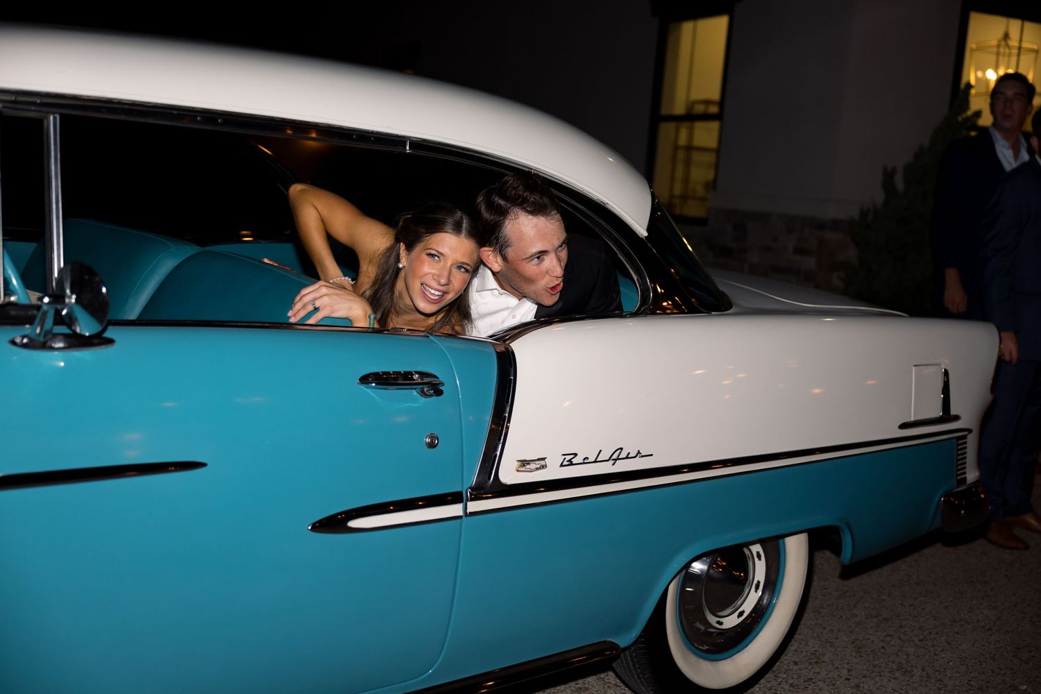 Couple leaning out of a classic teal and white 1950s Bel Air car at a nighttime event.
