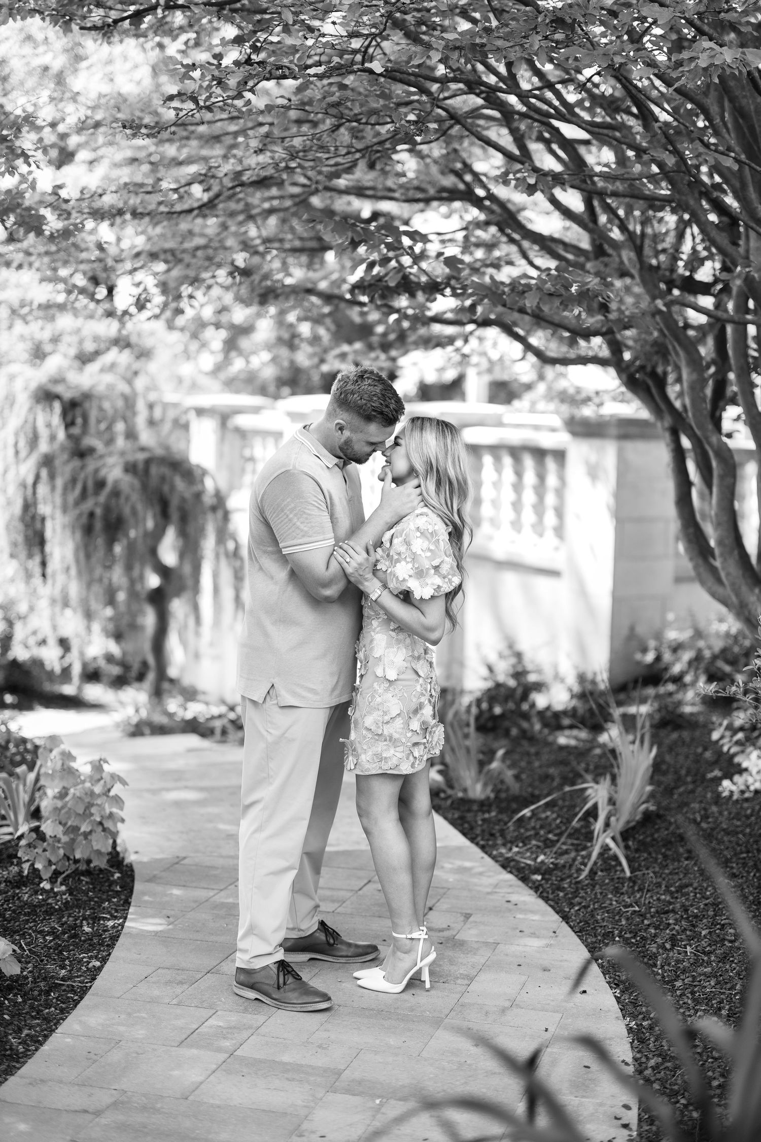 Black and white photo of a couple sharing a romantic moment on a garden pathway surrounded by lush trees and flowers.