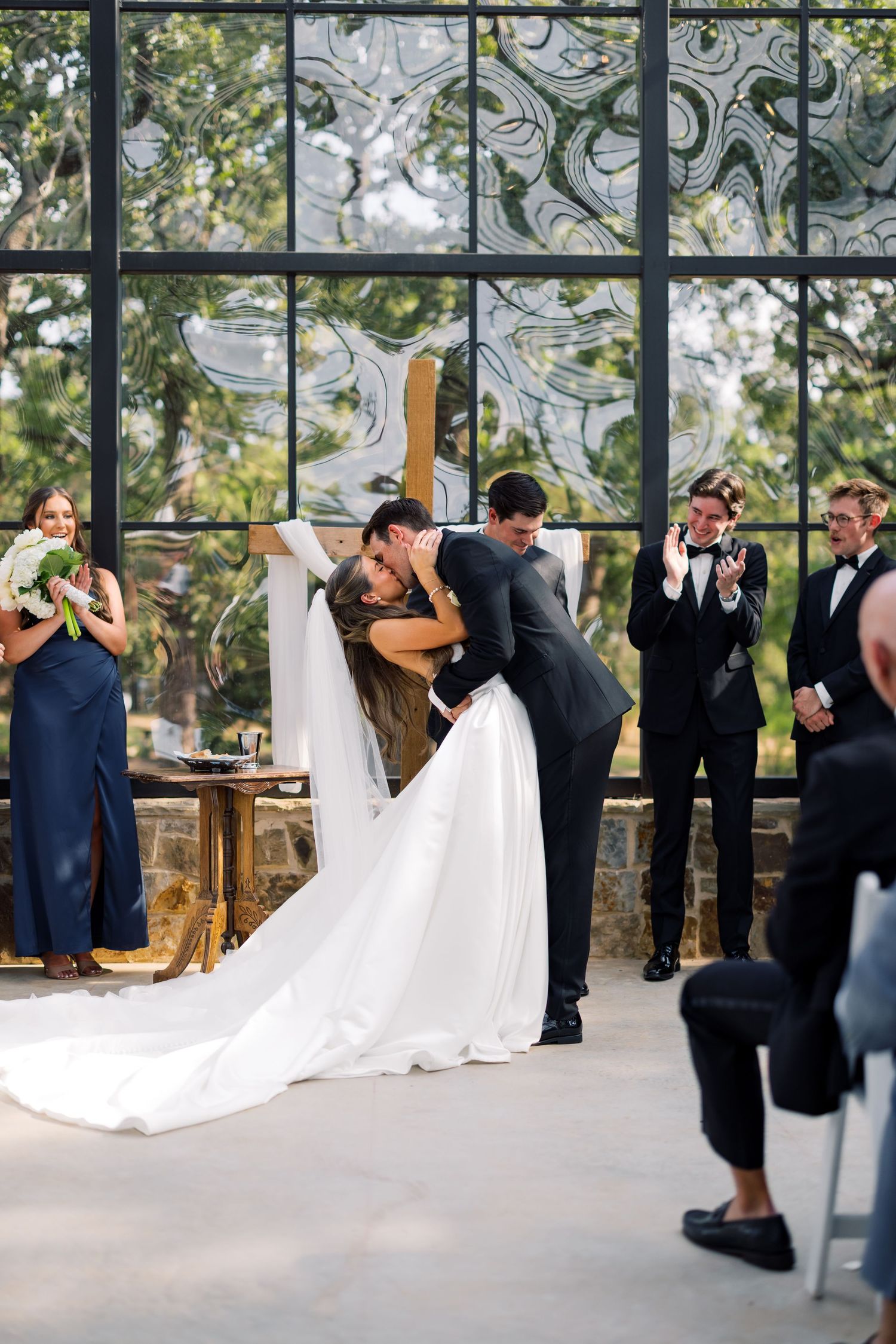 A groom dips his bride for a romantic kiss at the wedding altar, surrounded by cheering bridesmaids and groomsmen.