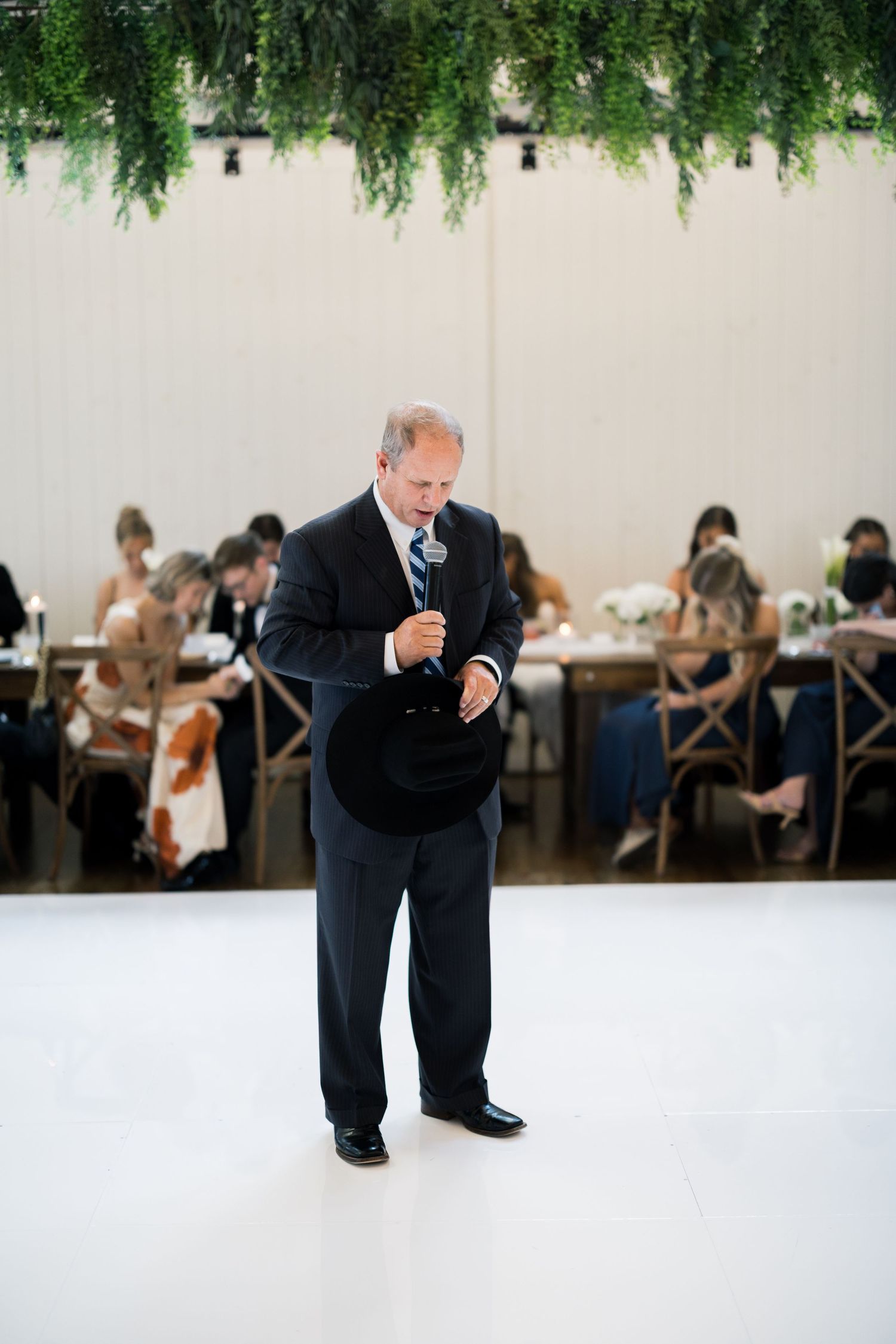 Man in dark suit giving a speech at an elegant wedding reception with greenery ceiling decor and guests seated at tables.
