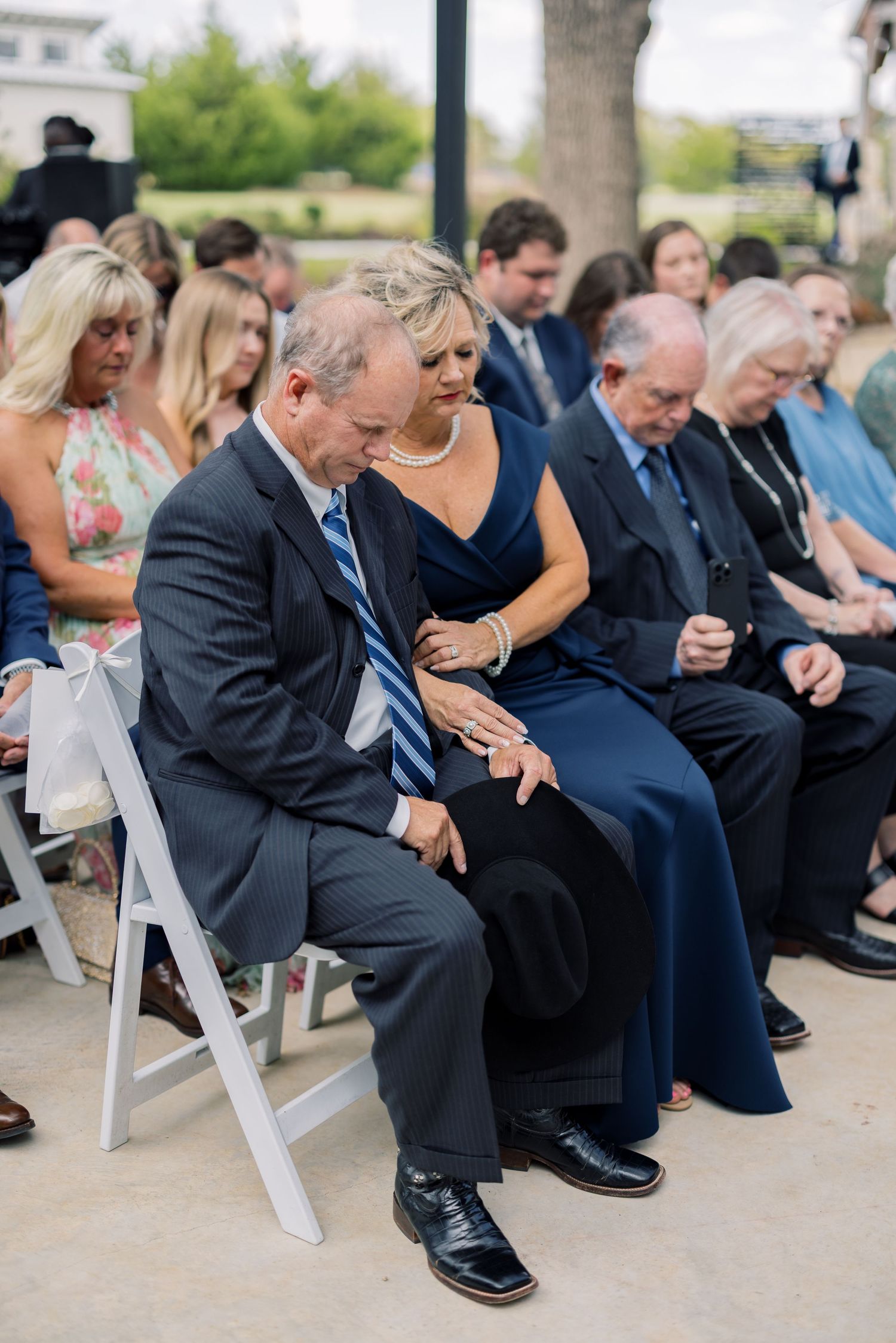Wedding guests in formal attire bow heads in prayer during an outdoor ceremony, seated on white chairs.