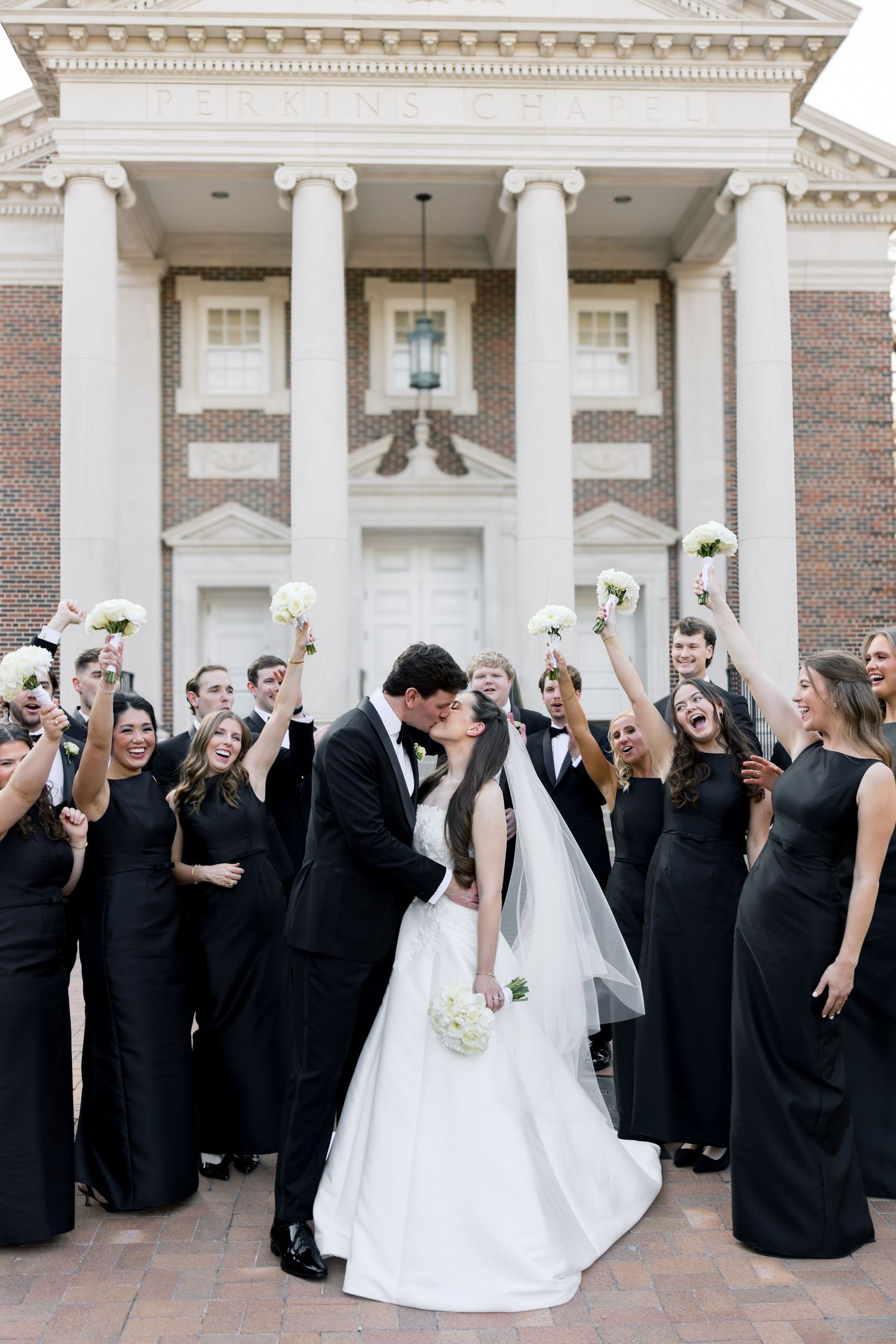 Wedding party in black attire cheering as bride and groom share a kiss before a brick building with white columns.