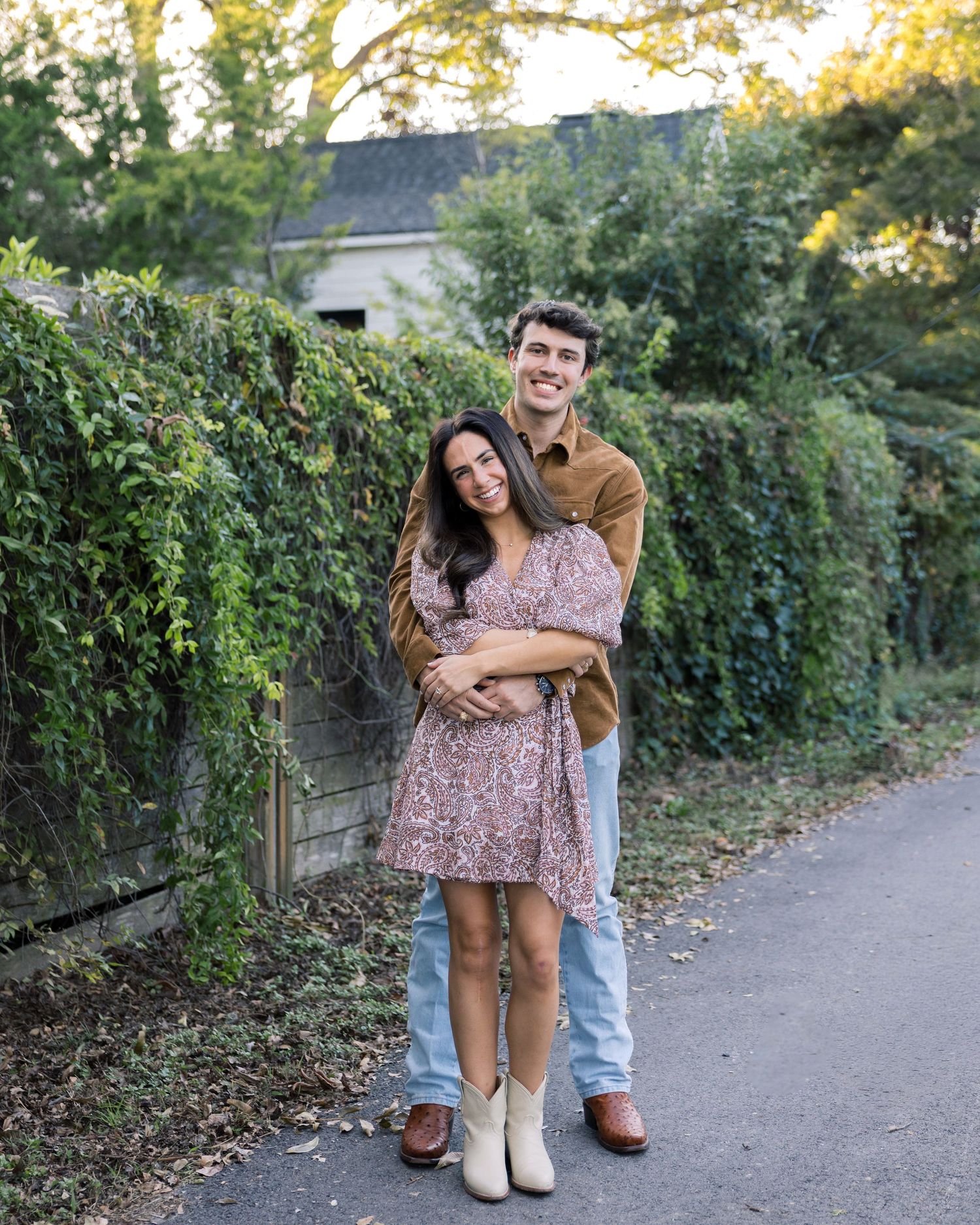 Couple embracing and smiling in front of an ivy-covered wall, woman in floral dress and white boots.