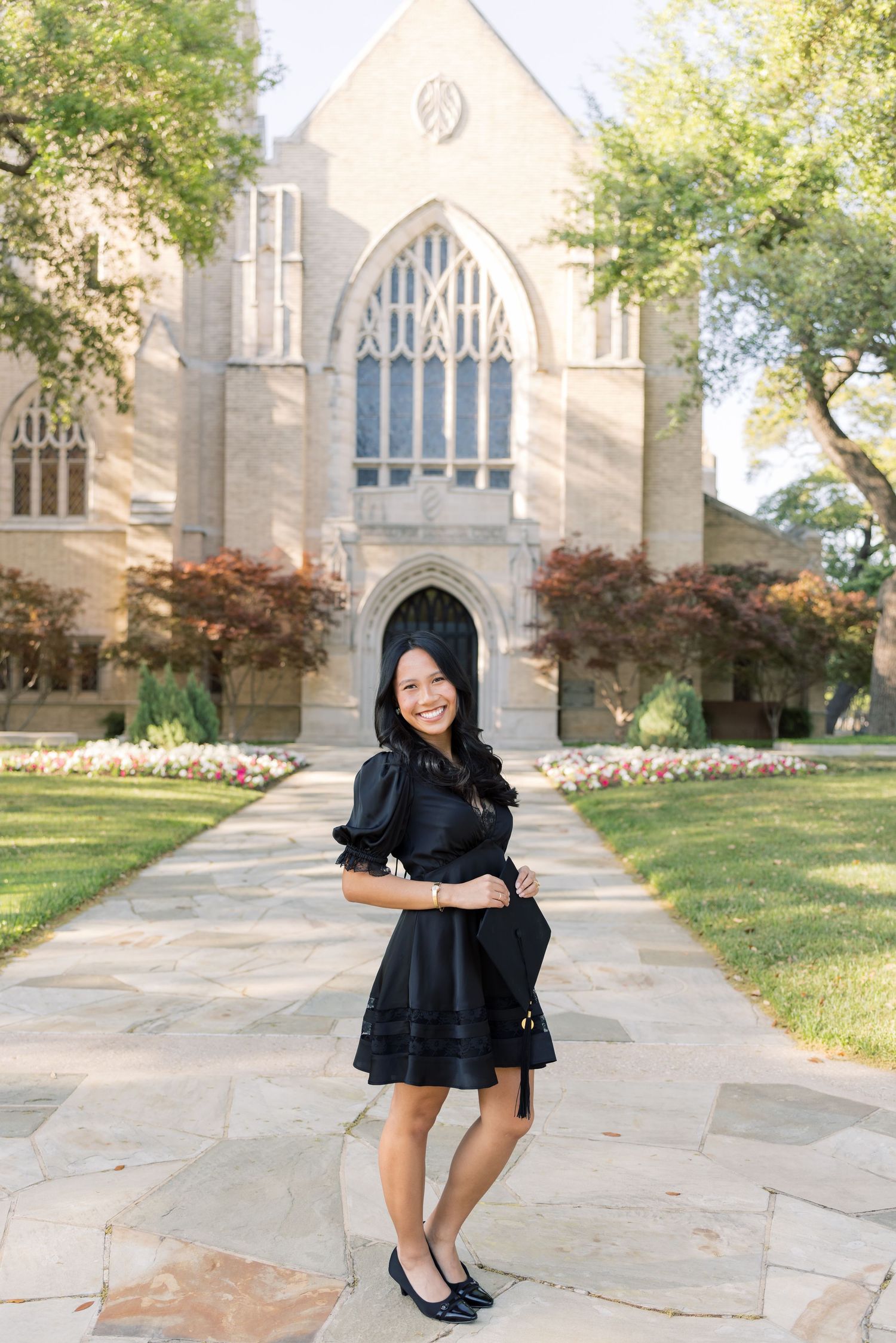 Smiling young woman in black dress poses on stone pathway before a Gothic-style university chapel.