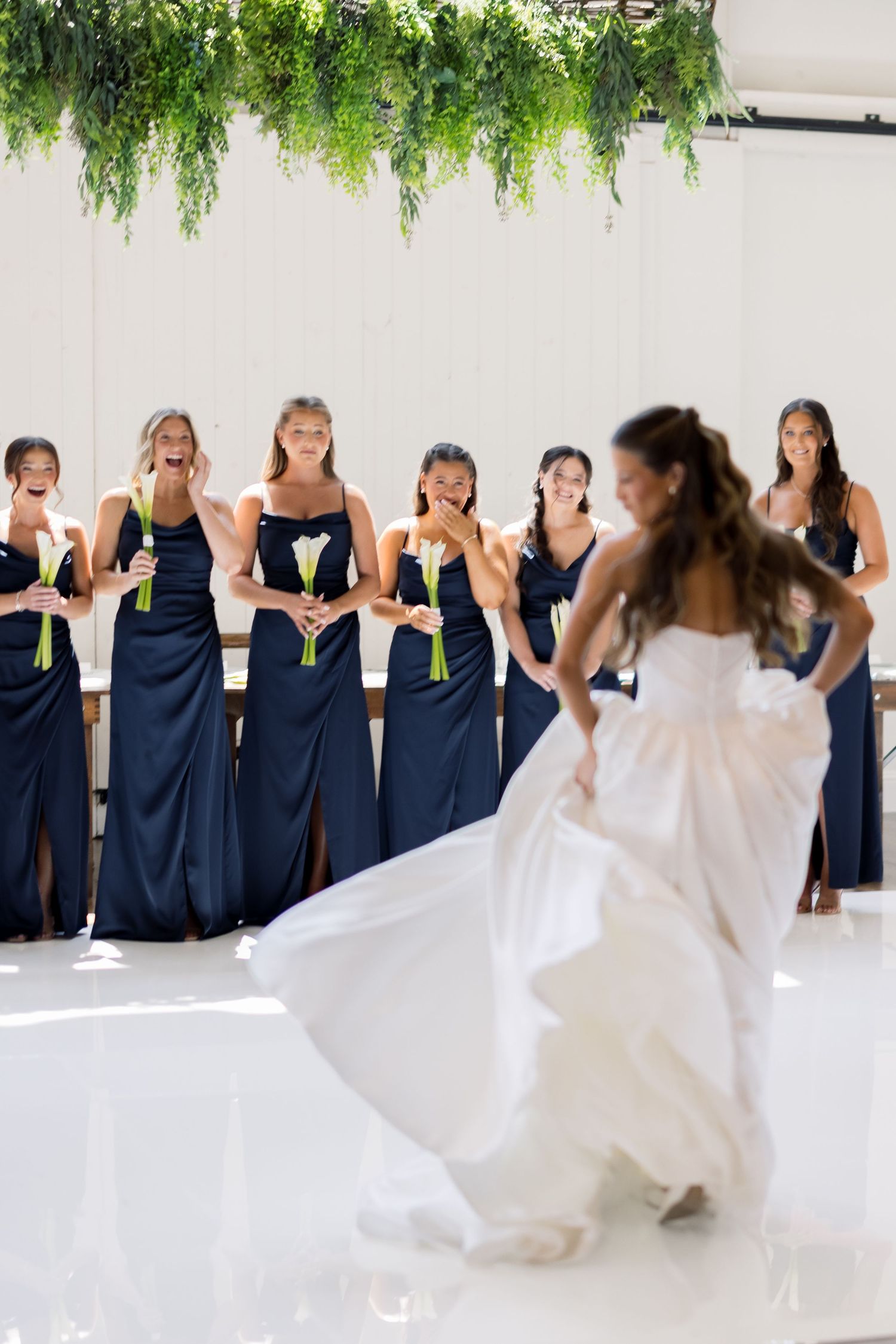 Bride in white gown twirls before bridesmaids in navy dresses holding white bouquets in elegant venue.