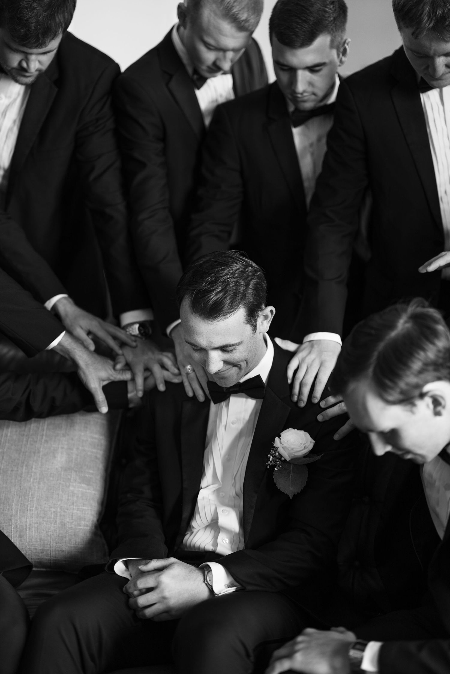 Black and white photo of a groom in a tuxedo laughing as groomsmen gather around him in celebration.