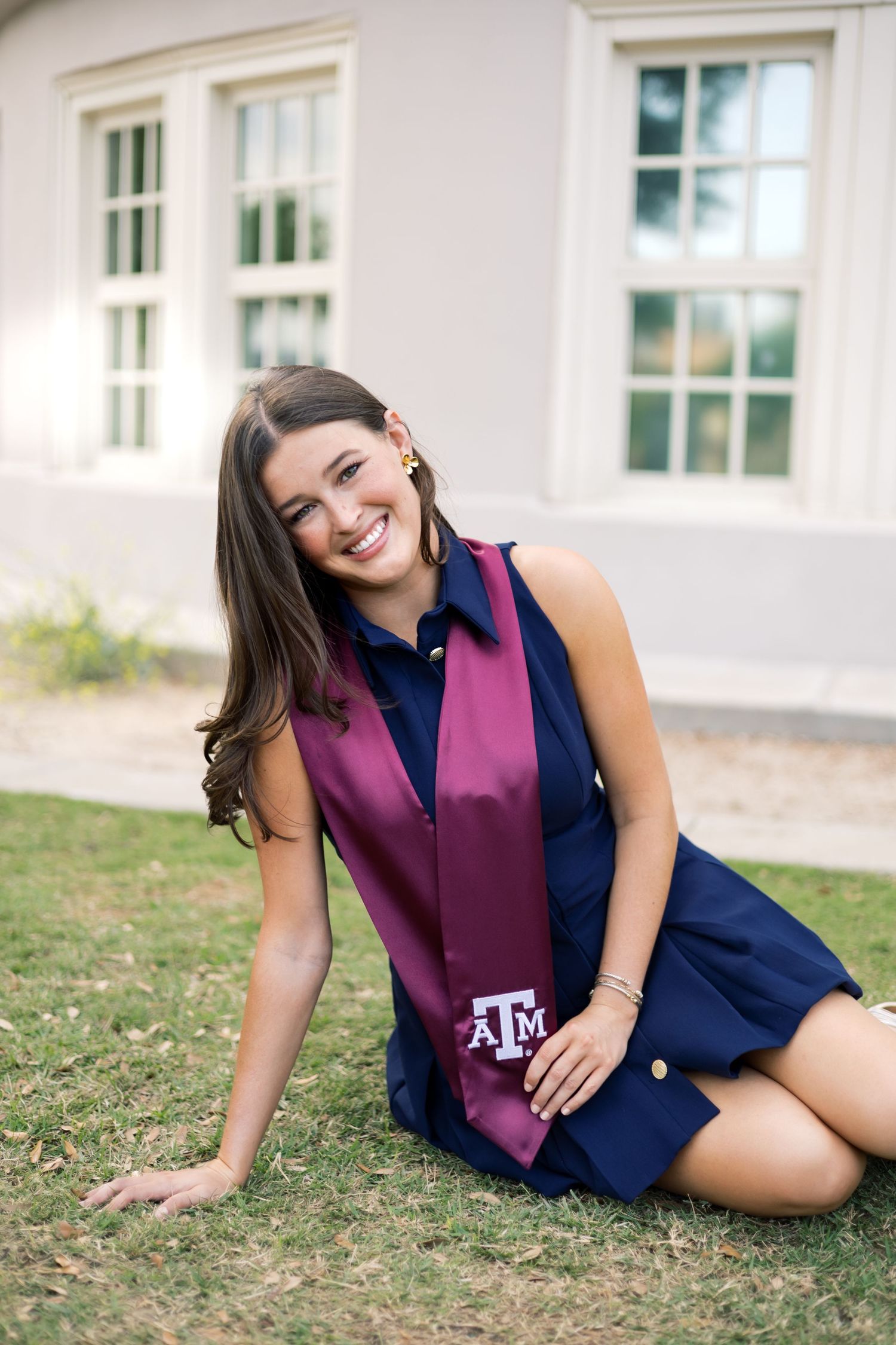 Happy Texas A&M senior sits on lawn wearing navy dress and maroon ATM graduation stole outdoors.