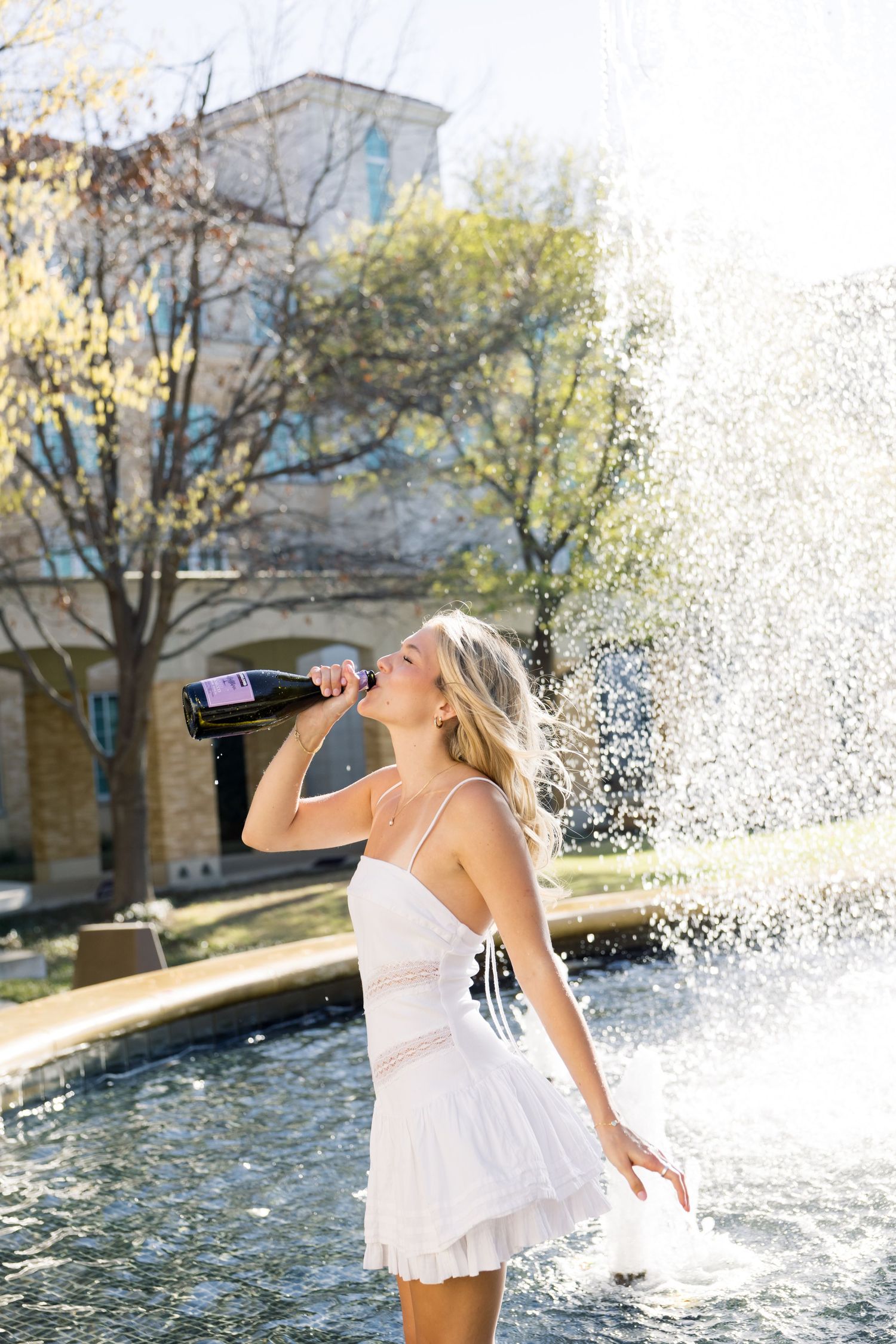 Blonde woman in white dress drinking from champagne bottle at sunlit campus fountain.