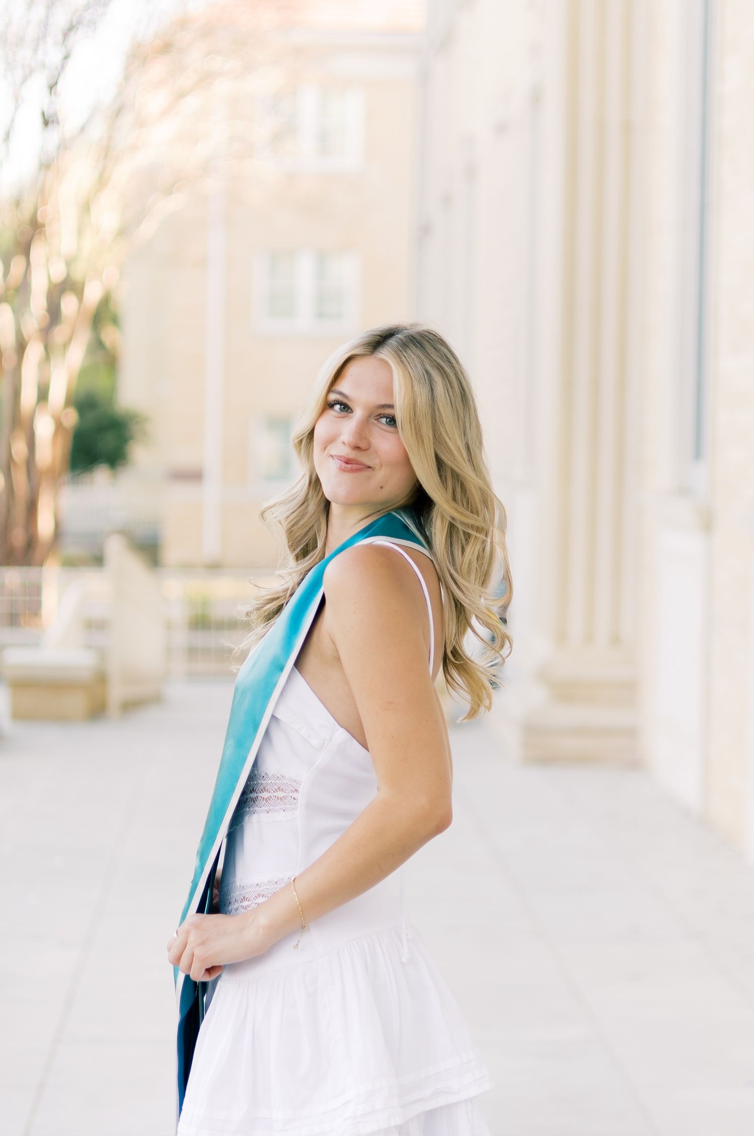 Blonde woman in white dress with teal graduation stole looking over shoulder at TCU campus.