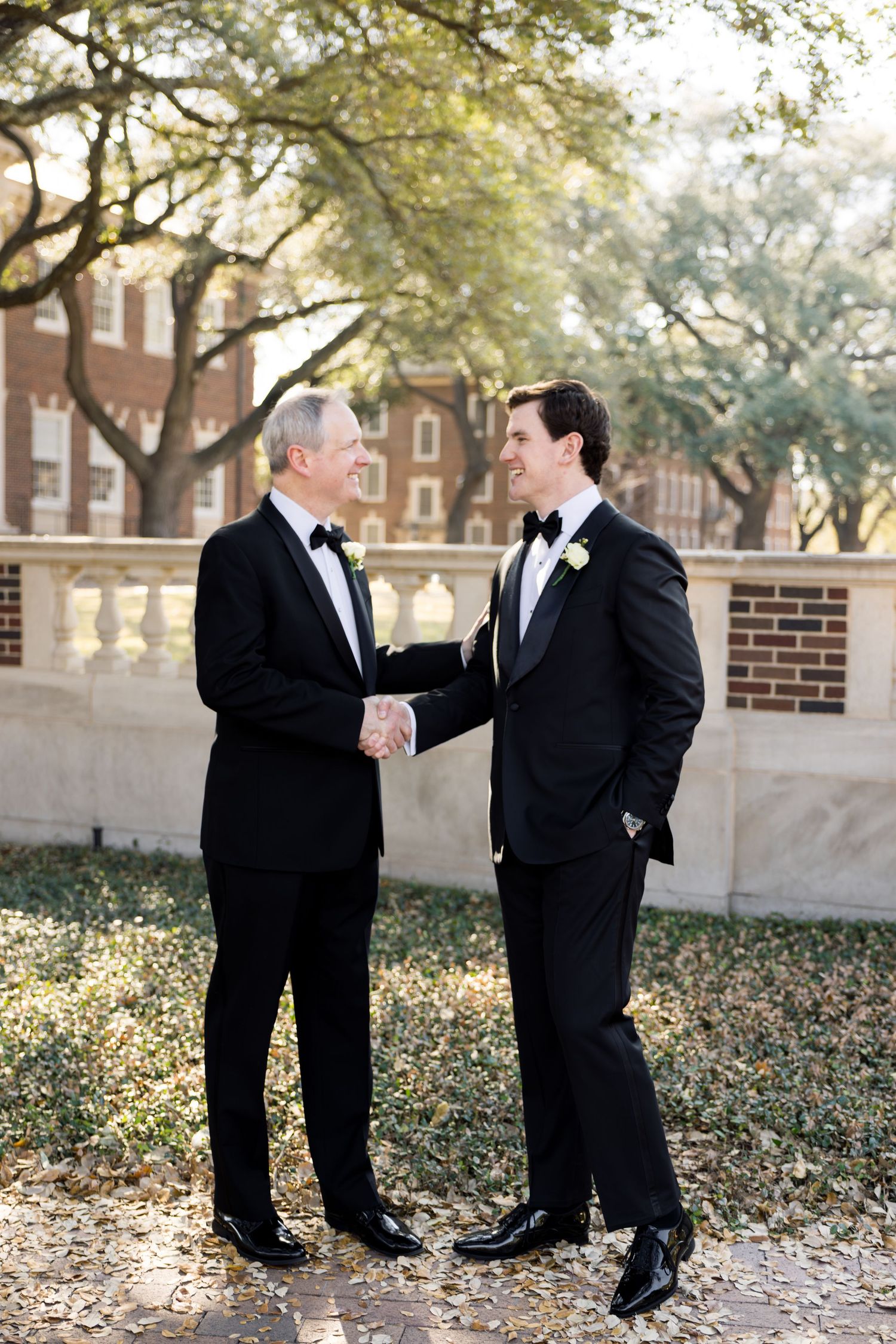 Groom and father of the bride share a handshake outdoors under oak trees on wedding day.