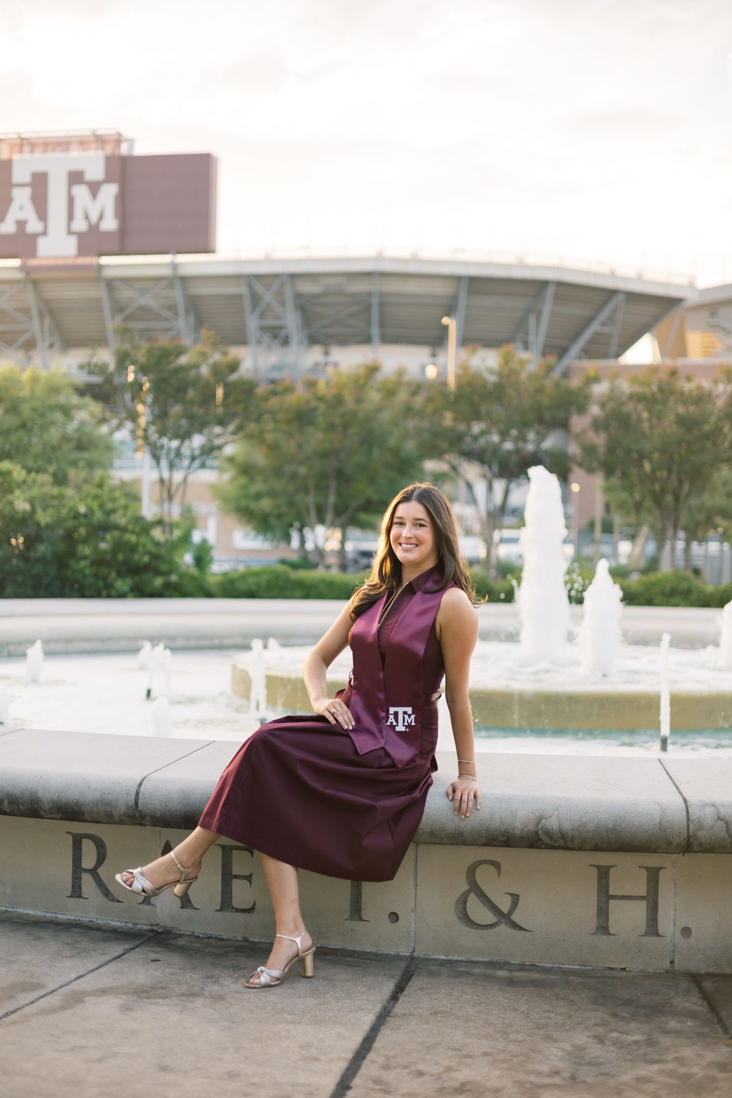 Texas A&M graduate in maroon dress sitting on fountain ledge with Kyle Field in background.