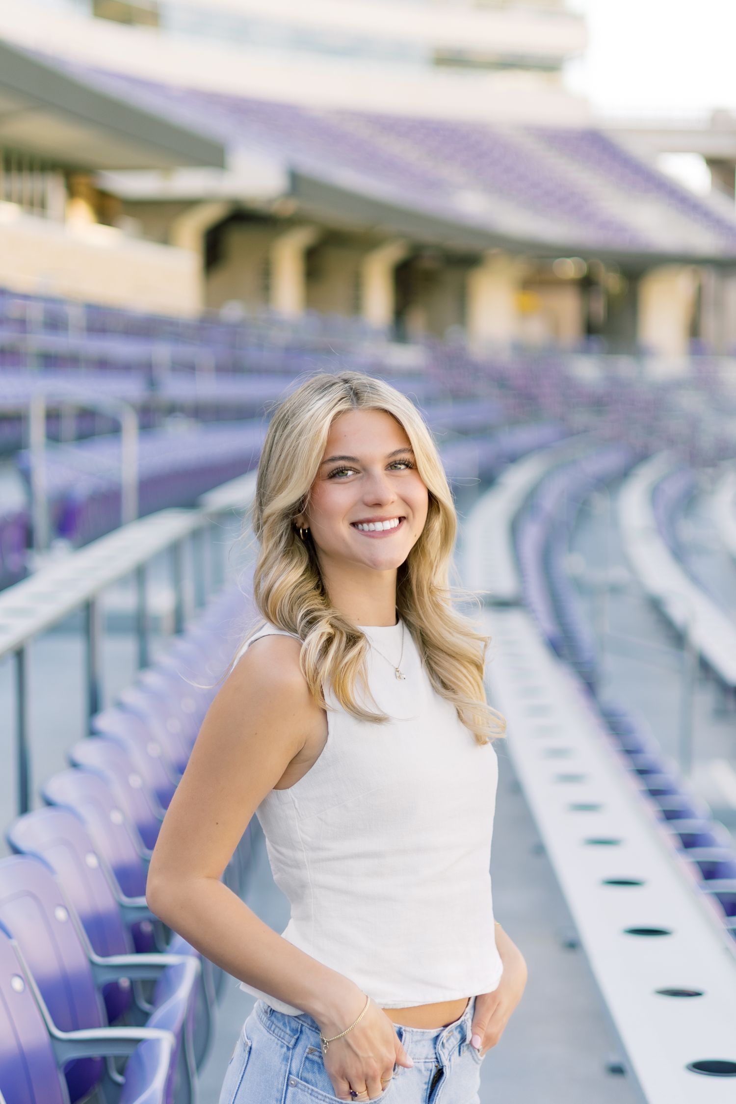 Smiling blonde woman in white crop top and jeans posing in empty purple stadium bleachers.