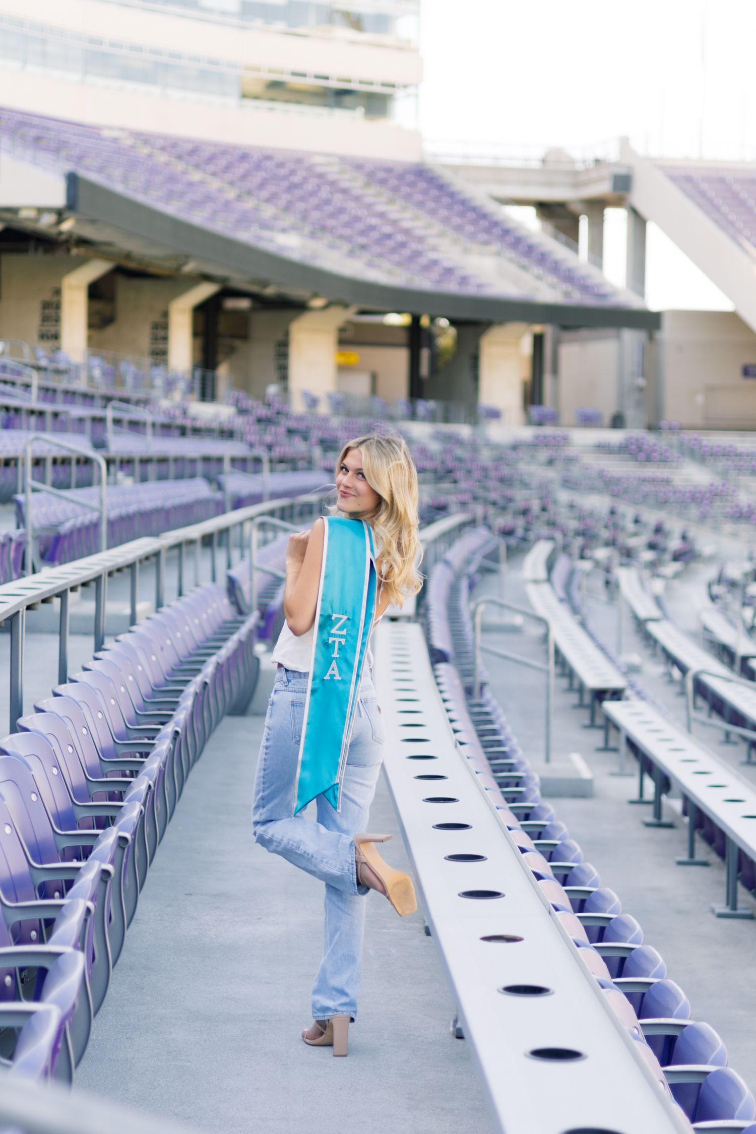 Blonde woman in jeans and white top walks through purple stadium seats wearing a teal ZTA sash.