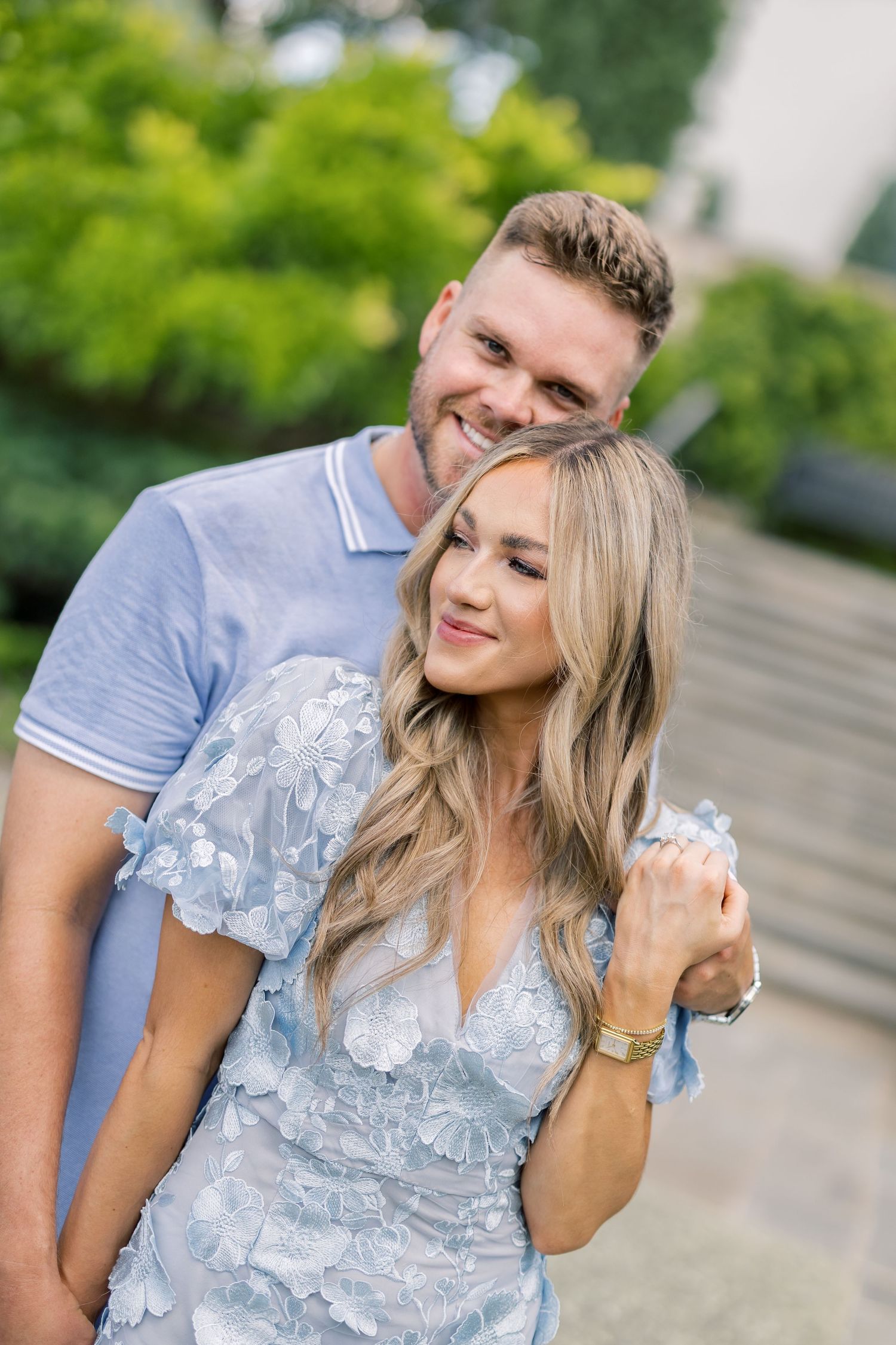 Smiling couple in matching blue outfits embracing outdoors, woman in floral lace dress, man in polo shirt.