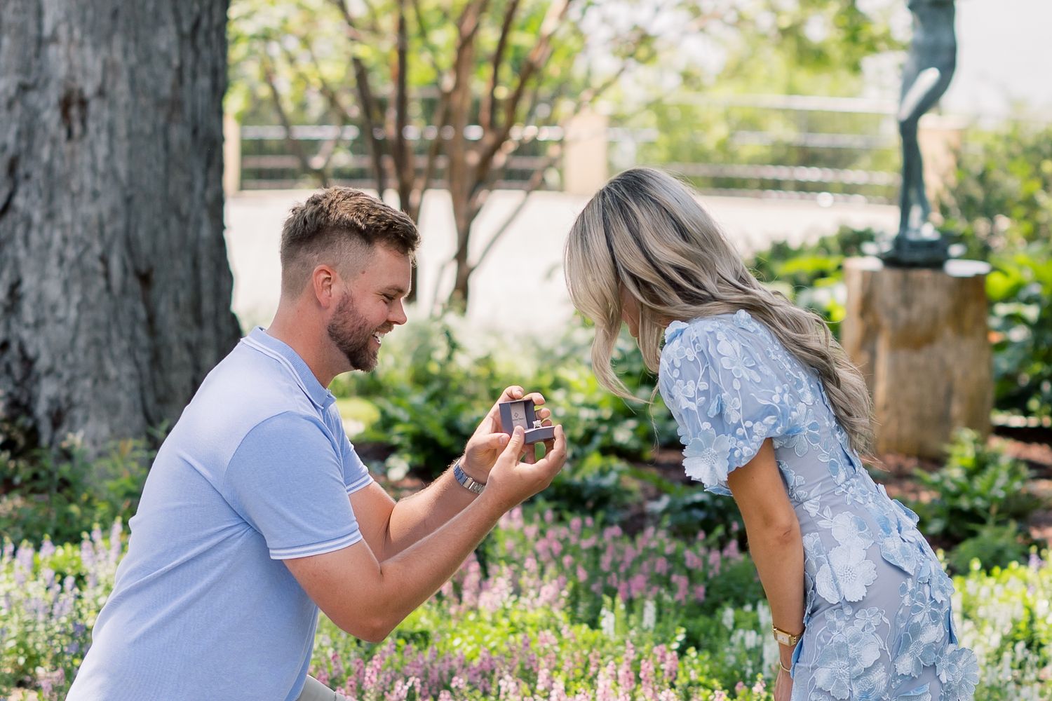 Man kneeling on one knee proposing with a ring box to a woman in a floral dress in a garden setting.