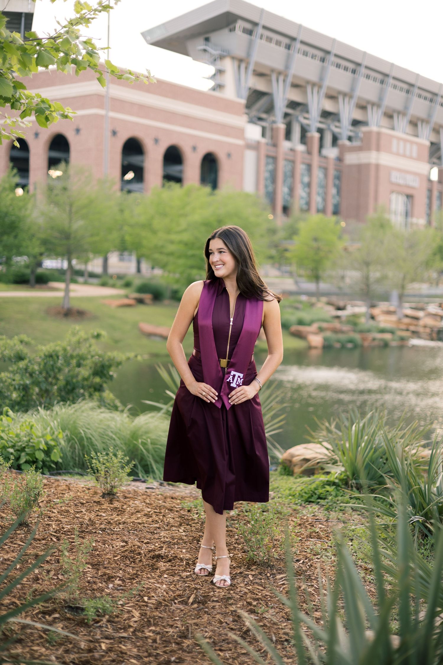Young woman in maroon dress with Texas A&M lanyard poses outdoors near Kyle Field stadium.