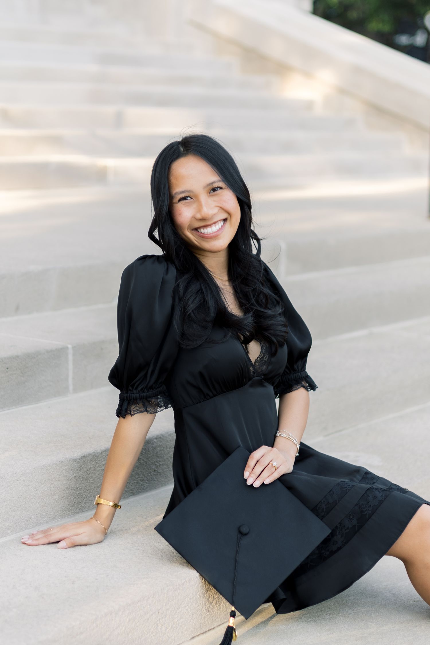 Smiling graduate in black dress sits on marble steps holding graduation cap at outdoor ceremony.