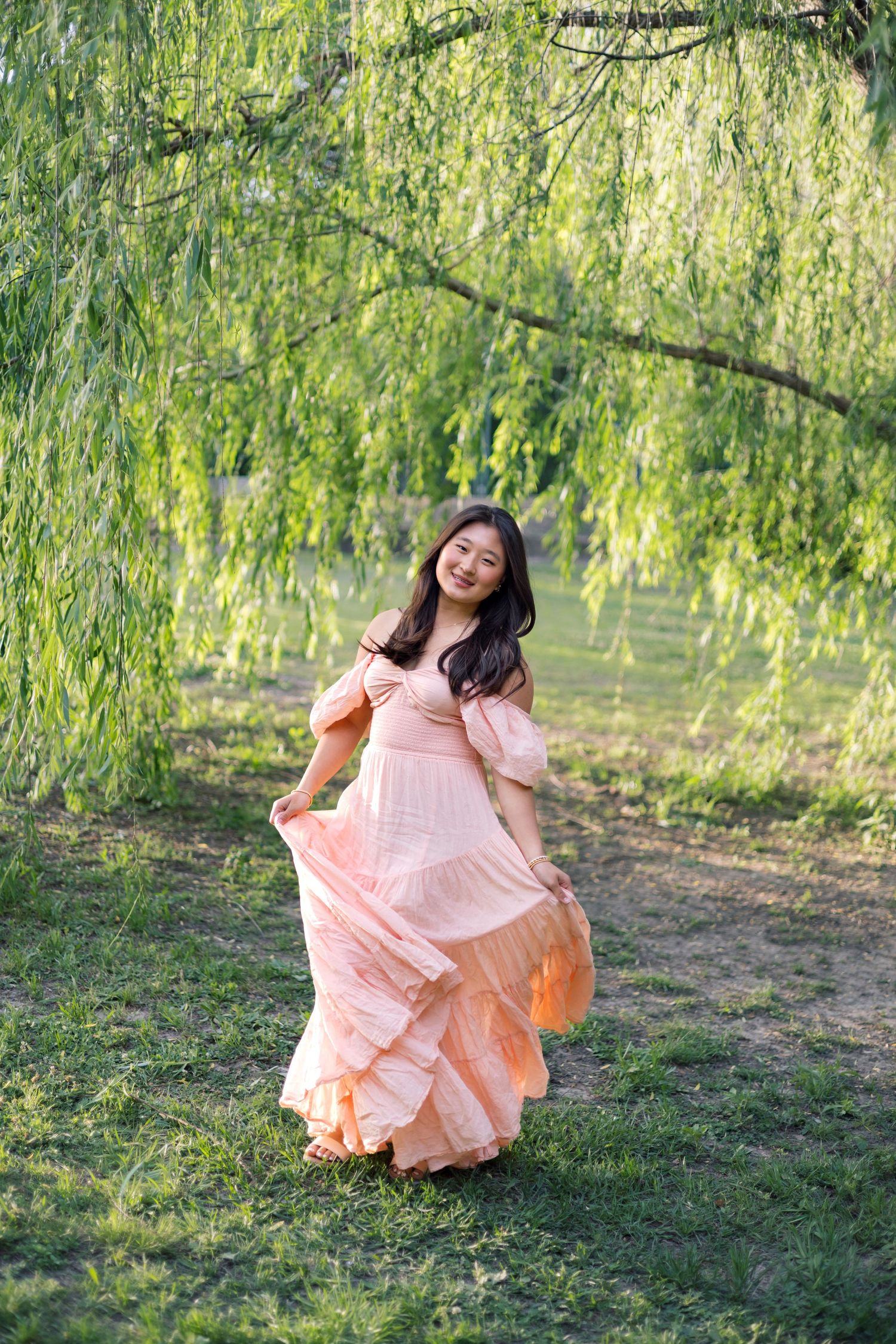 Young woman in flowing peach dress posing gracefully under weeping willow tree in green park.