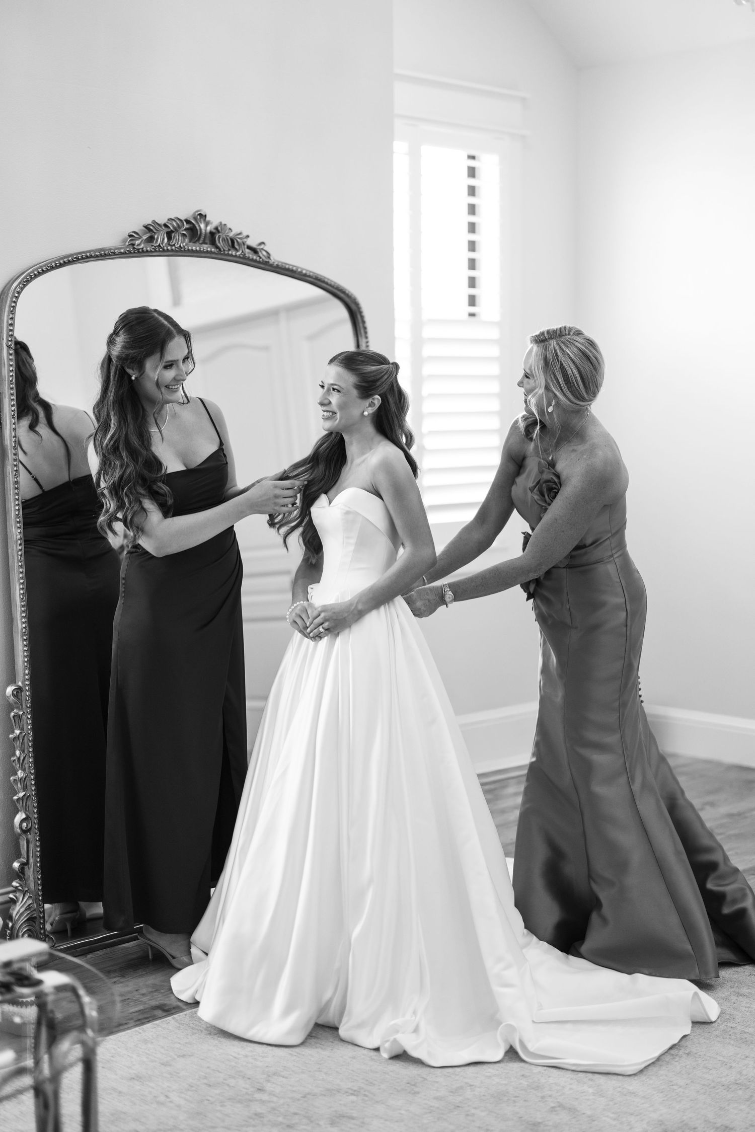 Bride in white gown assisted by bridesmaids near ornate mirror in black and white wedding photo.