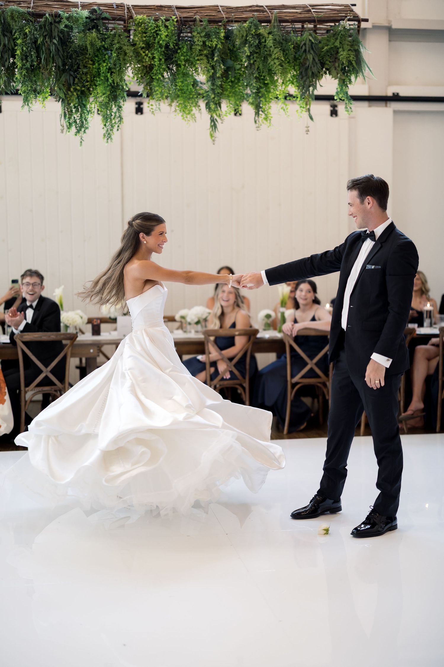 Bride in white gown twirls with groom in black tuxedo during first dance at elegant wedding reception.