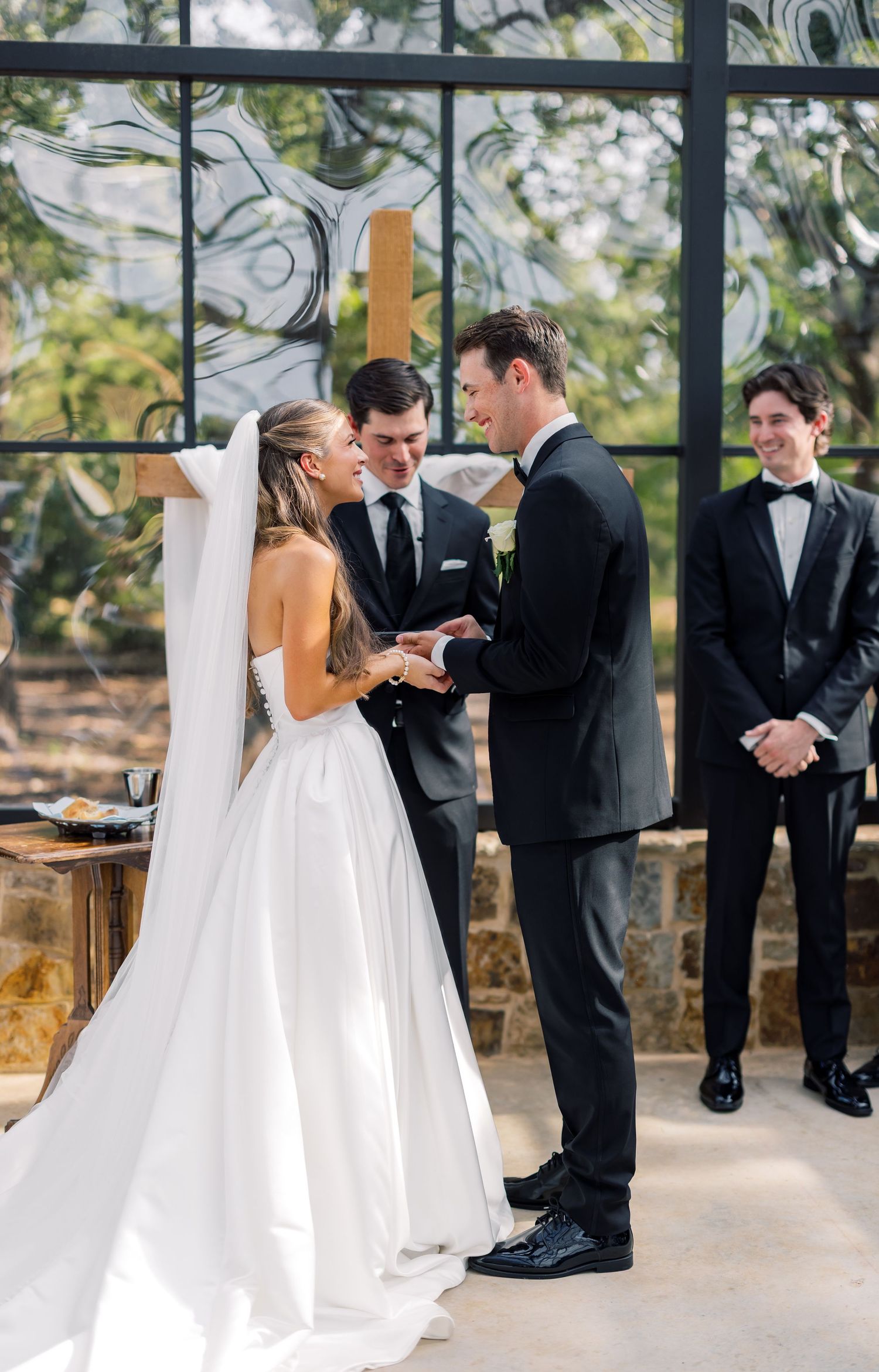 Bride and groom exchange rings at elegant wedding ceremony, officiant and groomsman smiling in background.