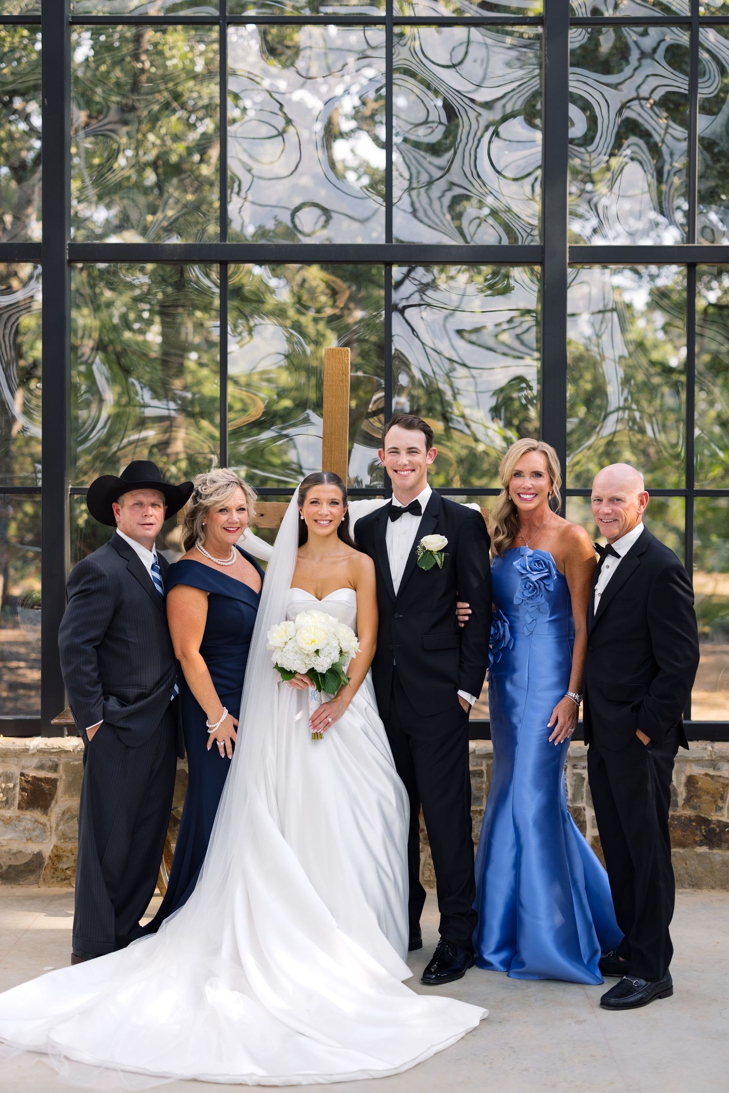 Wedding party of six posing before a glass chapel, bride in white gown holding bouquet, groom in tuxedo, guests in blue and black.