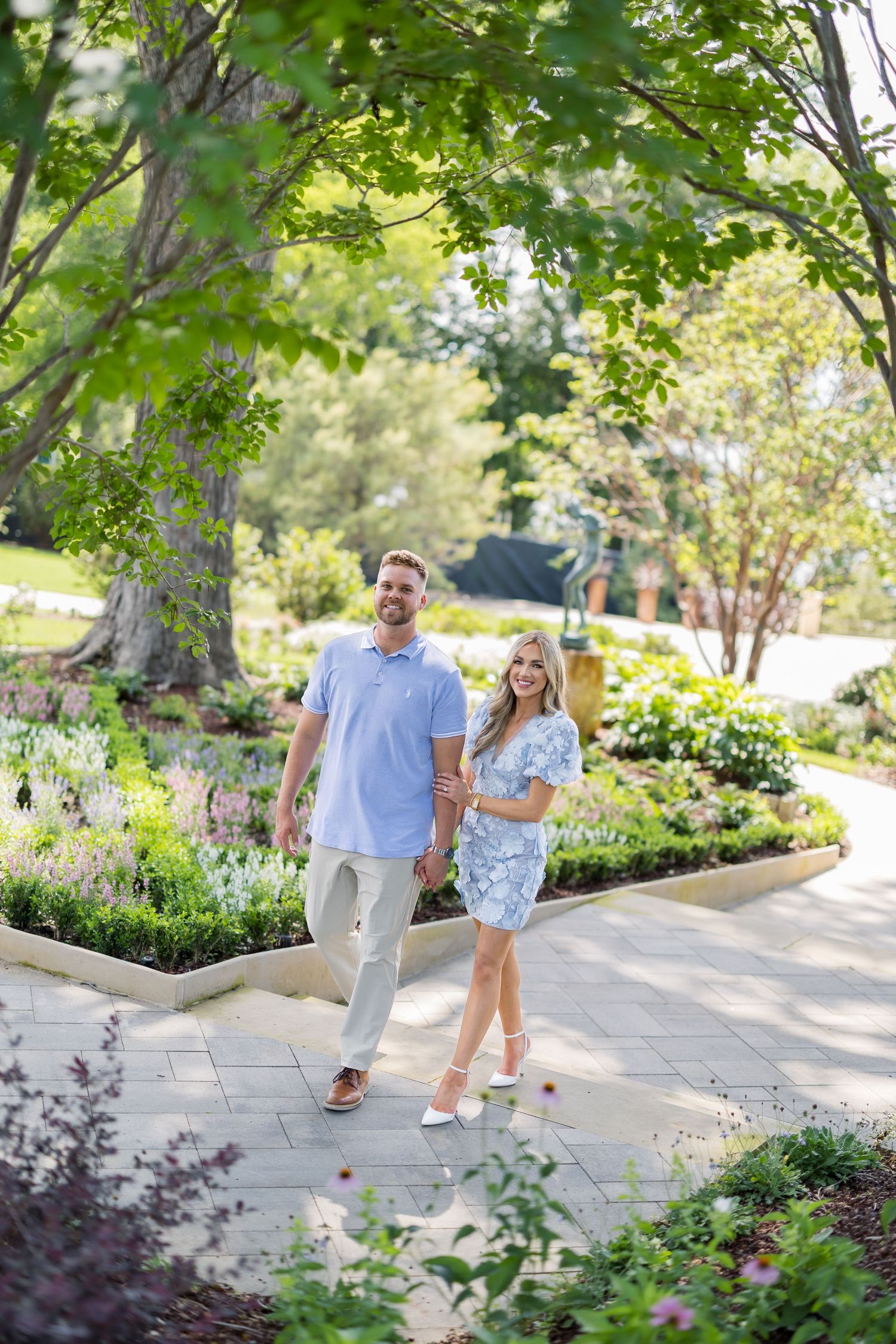 Couple holding hands and smiling while strolling through a lush garden path surrounded by colorful flowers and green trees.